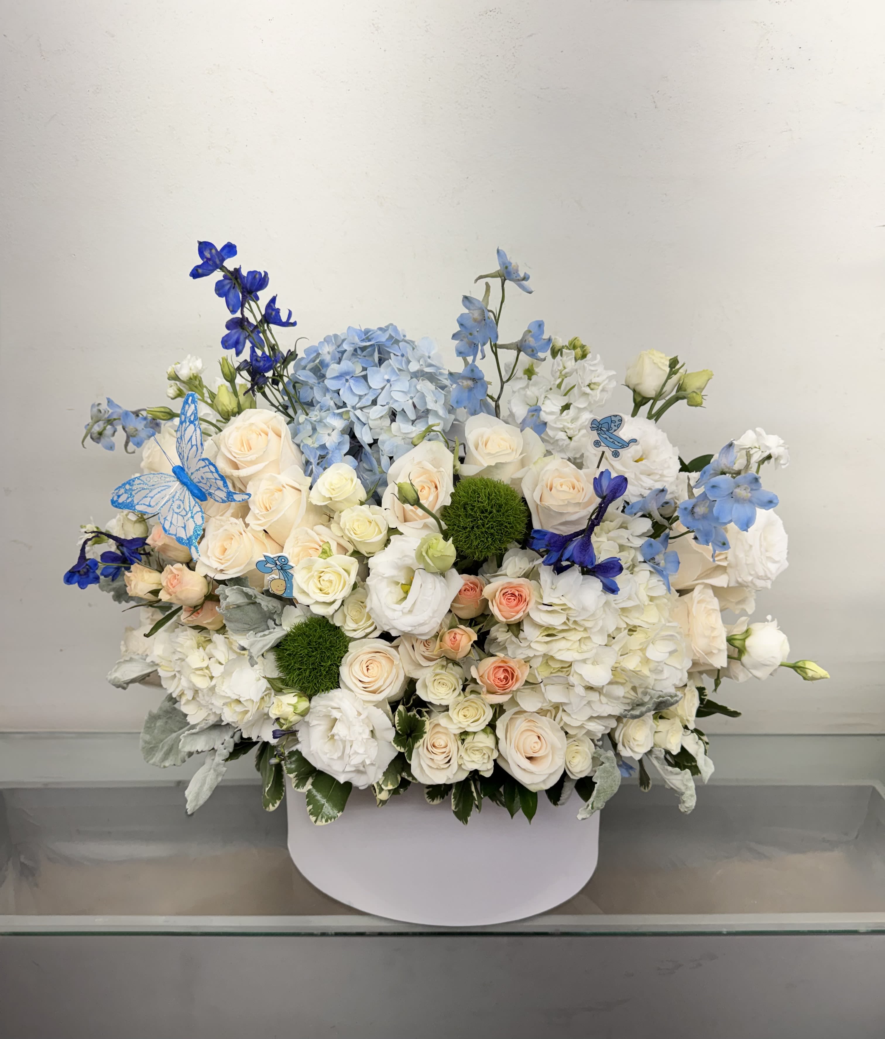 Blues Skies and Butterflies - Celebrate the arrival of a precious little one with our Baby Boy Garden Hatbox, a luxurious floral arrangement designed in soft, timeless shades of blue and ivory. This elegant keepsake arrangement features premium blue hydrangea, ivory roses, white lisianthus, delicate spray roses, blue delphinium, and lush seasonal blooms, beautifully arranged in a modern white hatbox.