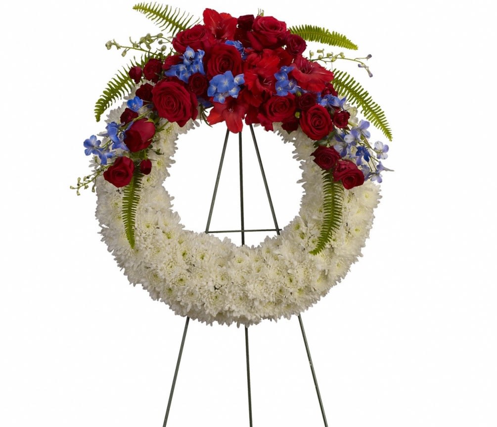 Circular funeral wreath with red roses, white flowers, and blue accents on a stand