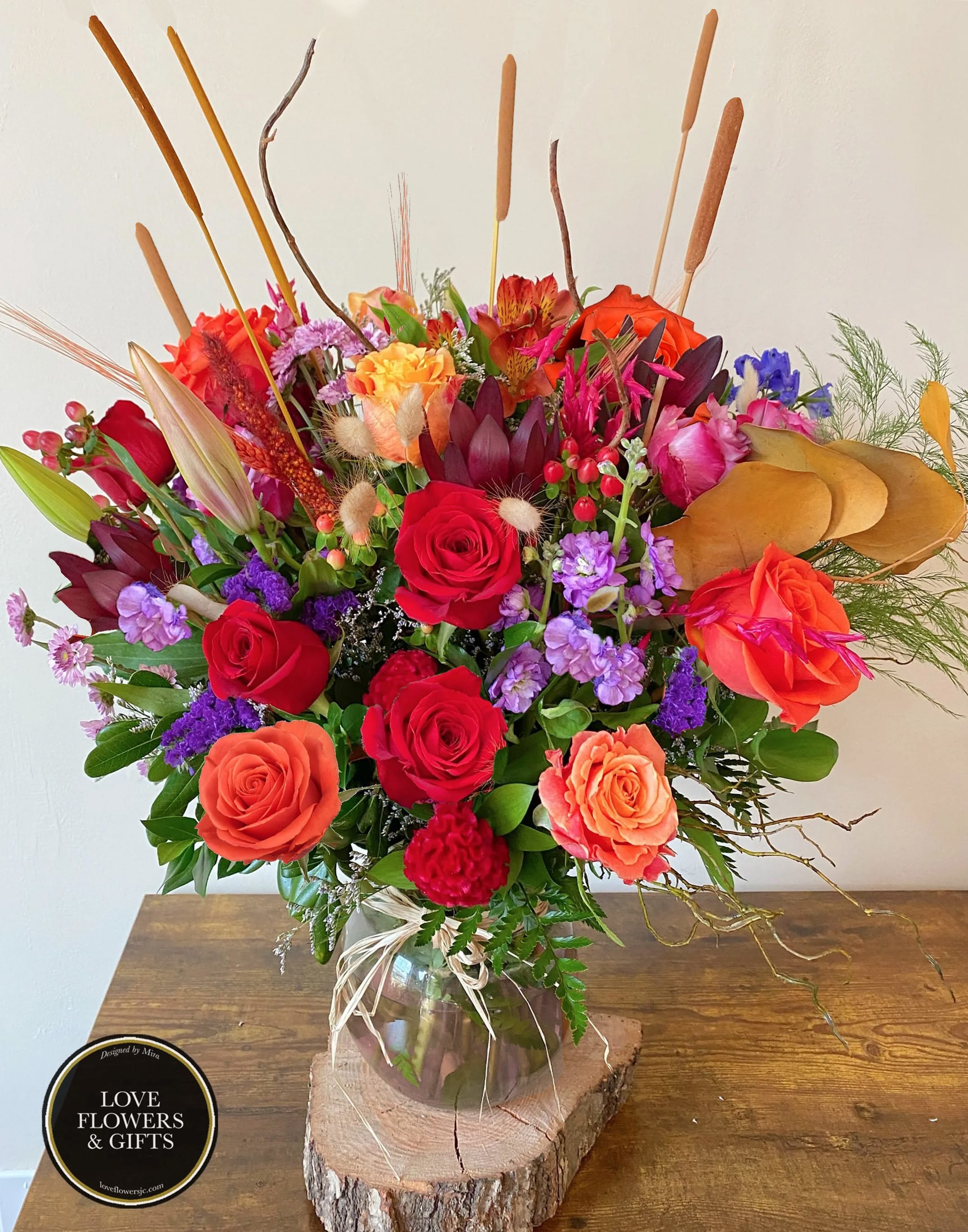 Tall mixed bouquet with red and orange roses, purple flowers, and cattails in a clear vase on a wood slice.
