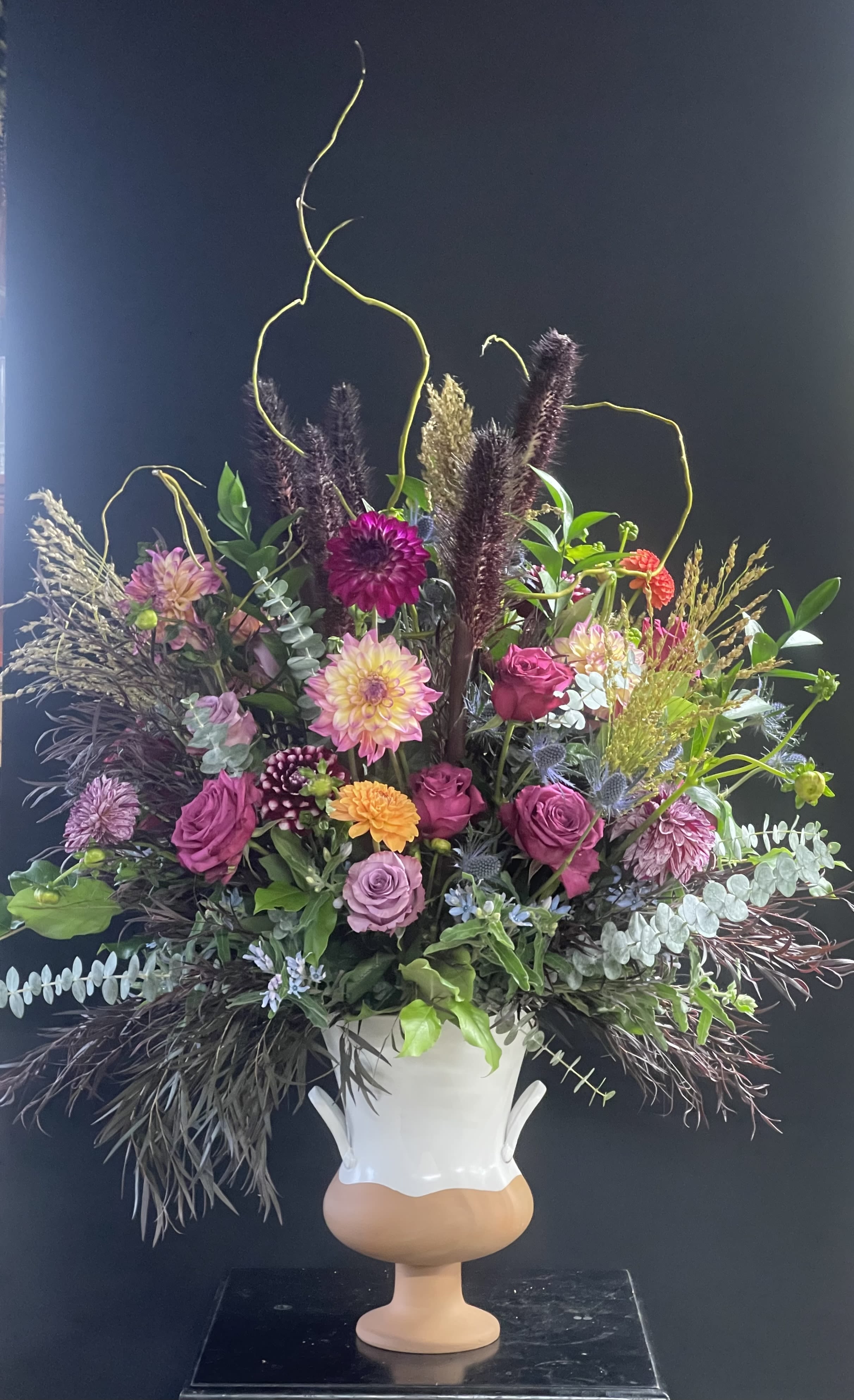 Mystic Willow Floral Arrangement - An enchanting blend of moody elegance and wild beauty, this grand arrangement weaves together the velvety richness of dahlias and lavender roses, the unique textures of black millet, broom corn, and agonis, and the airy grace of blue tweedia. Baby eucalyptus, salal, and curly willow add depth and movement, evoking the feeling of a windswept woodland at dusk. Perfect for making a bold, artistic statement in any space.