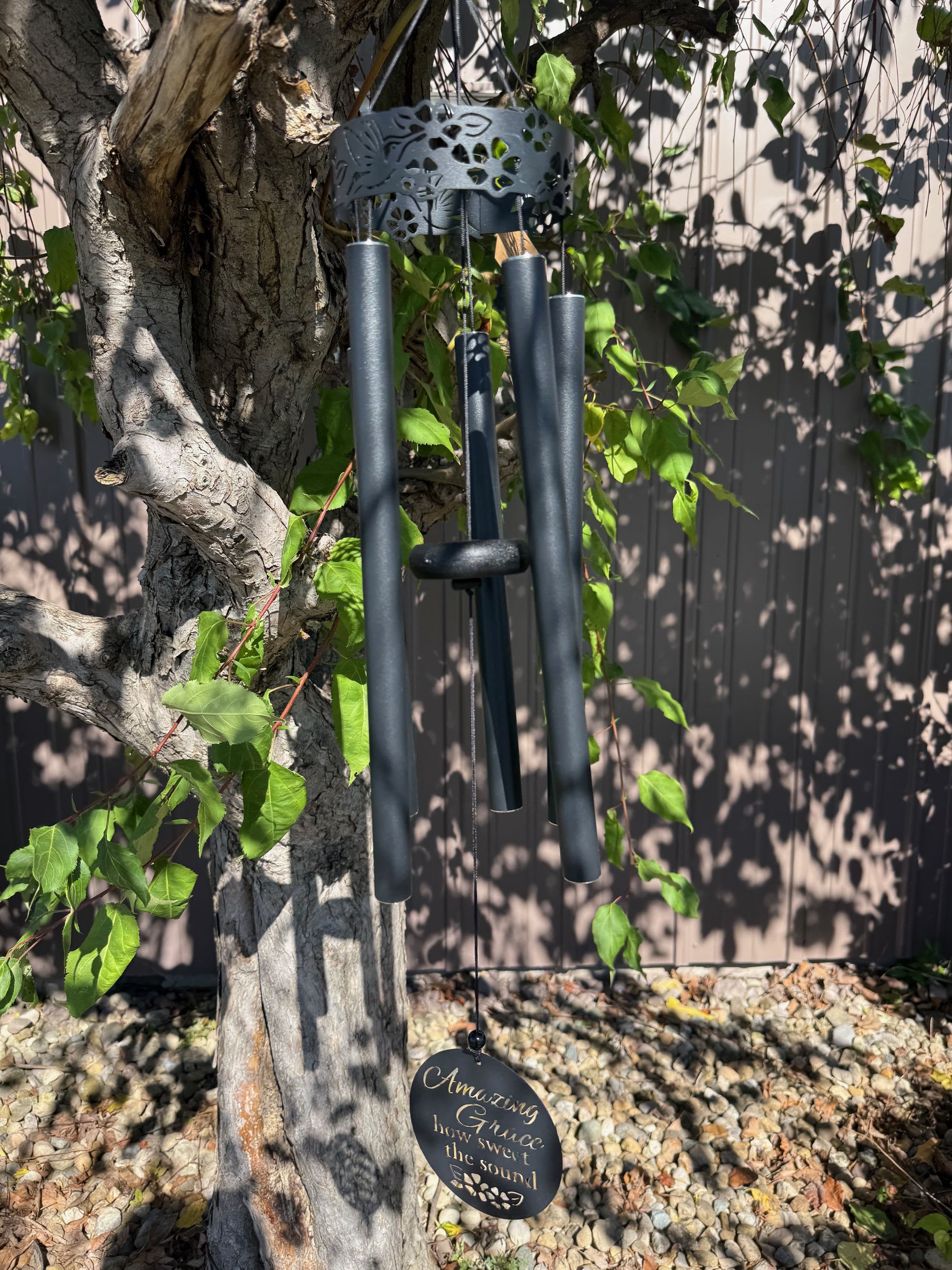 Black metal wind chime with Amazing Grace inscription hanging from a tree outdoors