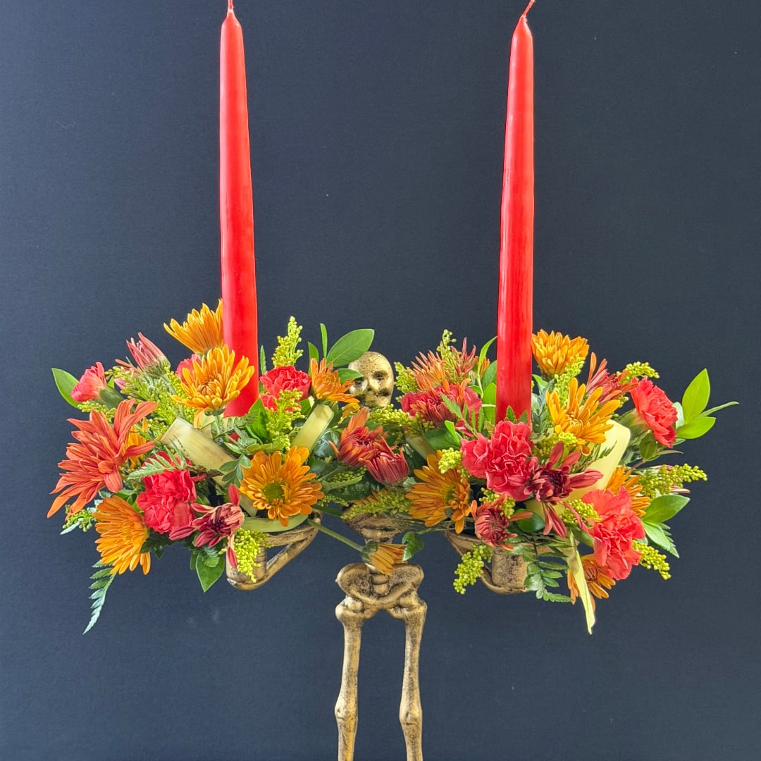 Two red taper candles rise from a colorful floral centerpiece with a skeleton base.