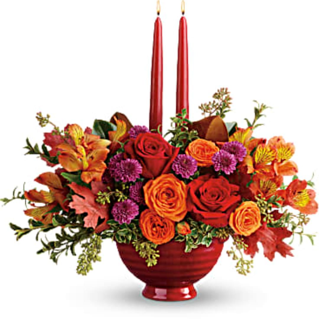 Autumn floral arrangement with red and orange roses in a red vase, accented by tall red candles.
