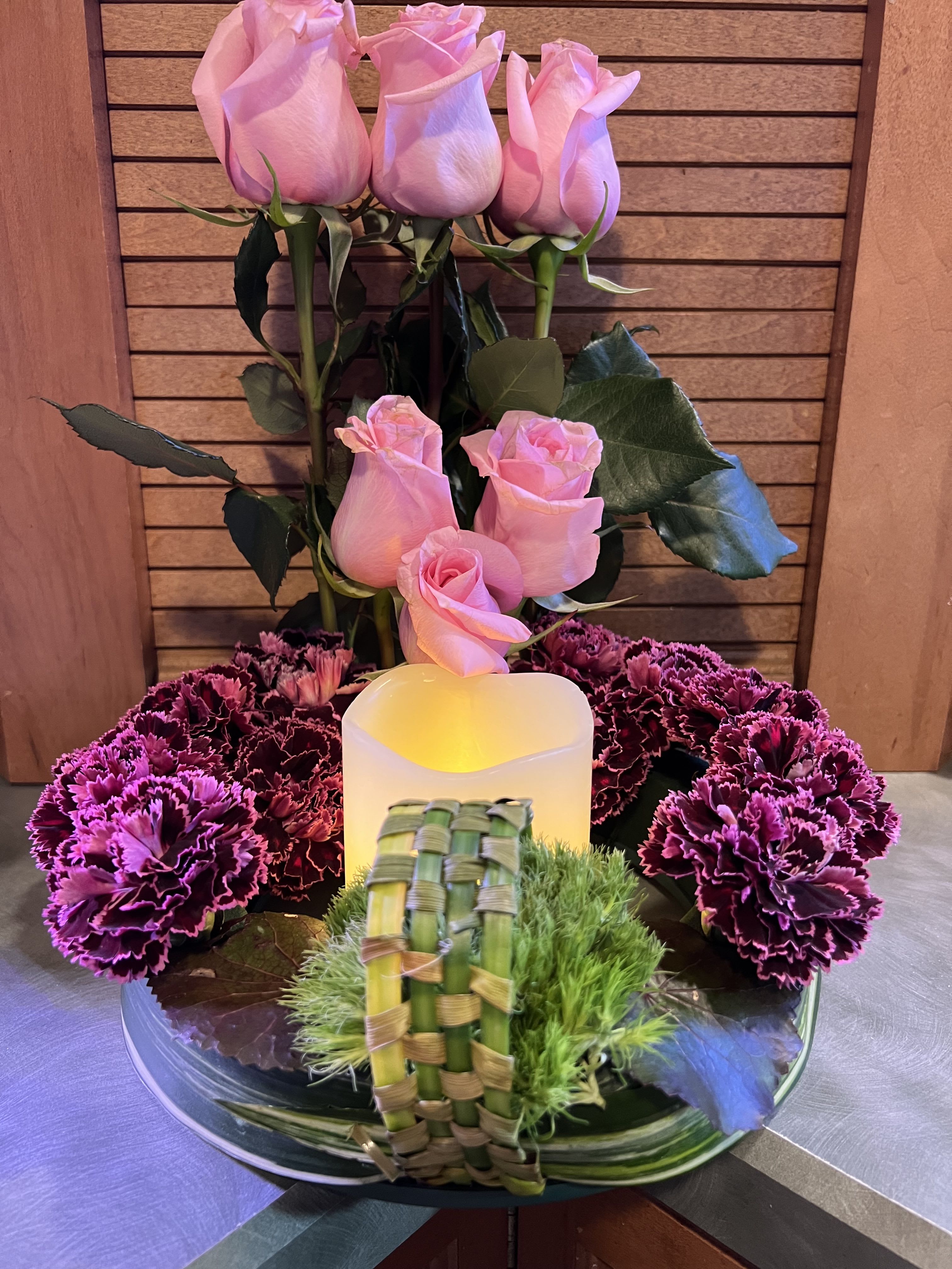 Peace of Mind  - Tall pink roses rise behind a low ring of ruffled purple carnations in this Peace of Mind flower arrangement, creating a layered, compact centerpiece perfect for a sympathy gesture, a romantic surprise, or a calming everyday gift. The soft white and green accents keep the palette soothing while the battery-operated candle in the center of the designer ring  casts a gentle glow, so the person you're thinking of will feel comforted and seen.  Arranged in a designer's ring with a woven grass accent, the dense circular base of white and green filler creates a handcrafted, decorative feel that suits a small celebratory table display or bedside table. The mix of roses and carnations gives both graceful height and plush texture; colors are pink, purple, white, and green as shown. The included battery candle provides safe, long-lasting ambiance without flame; the compact size makes it easy to place in tight spaces.  Choose this arrangement when you want to offer calm, warmth, and a quietly elegant impression-the kind of gift that makes someone feel genuinely cared for. For longevity, keep the floral foam hydrated as directed and replace the candle battery when needed; this piece arrives ready to display.