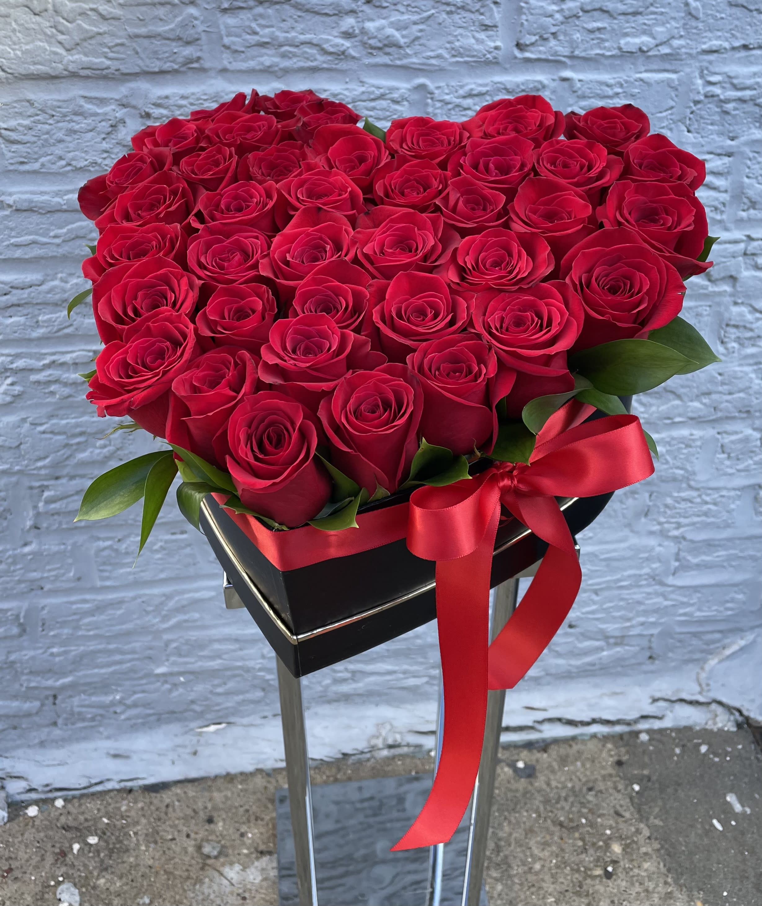 Heart-shaped arrangement of red roses with a red ribbon on a black stand