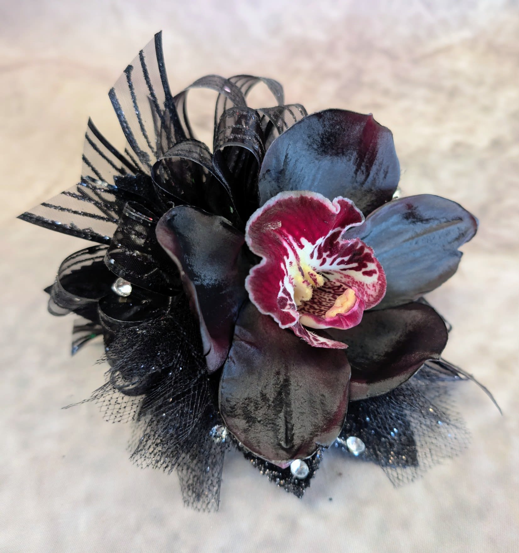 Noir Orchid Corsage With Rhinestone Accents - Make an unforgettable statement with our Black Orchid Corsage, a striking blend of sophistication and edge, exclusively from Windmill Floral Expressions in Las Vegas. This design showcases a dramatic black orchid with deep, velvety petals, highlighted by a vivid center of golden yellow and burgundy tones. Accented with shimmering black ribbon, sparkling crystals, and delicate textural details, it’s the perfect accessory for those who want to stand out with elegance and artistry.