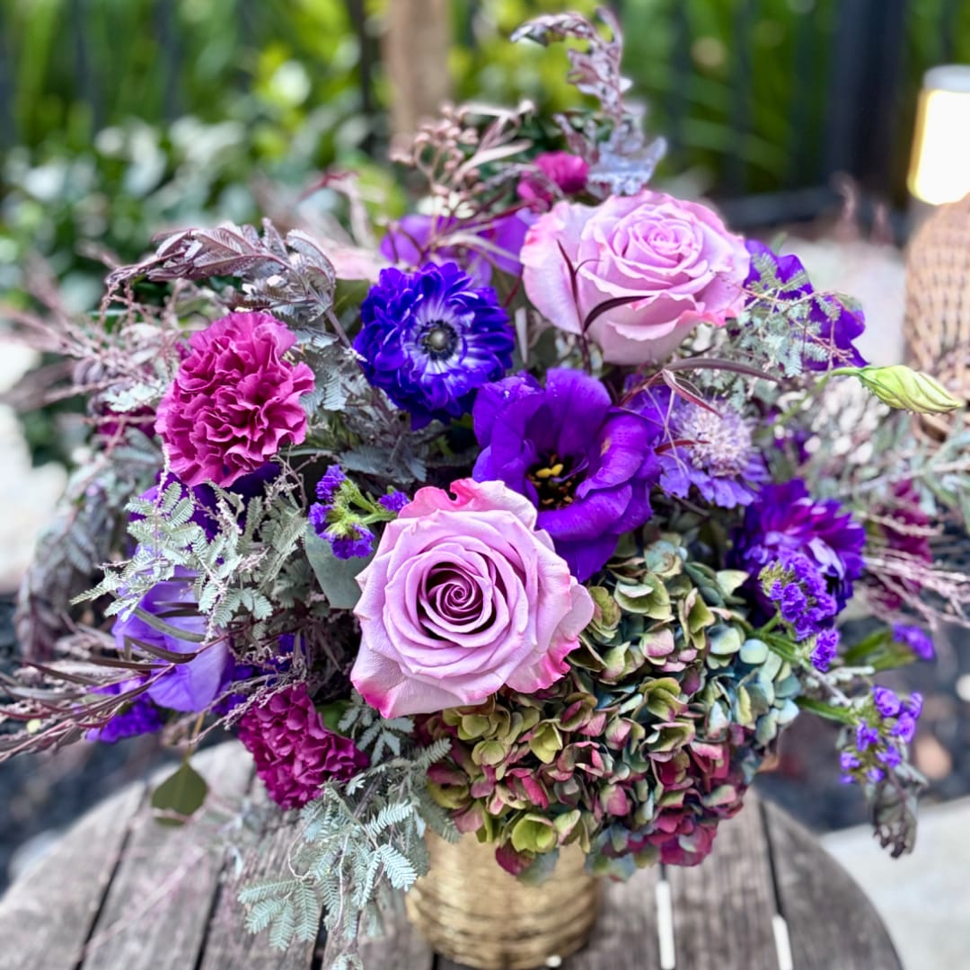 Purple and pink floral arrangement with roses, carnations, and hydrangeas in a gold vase