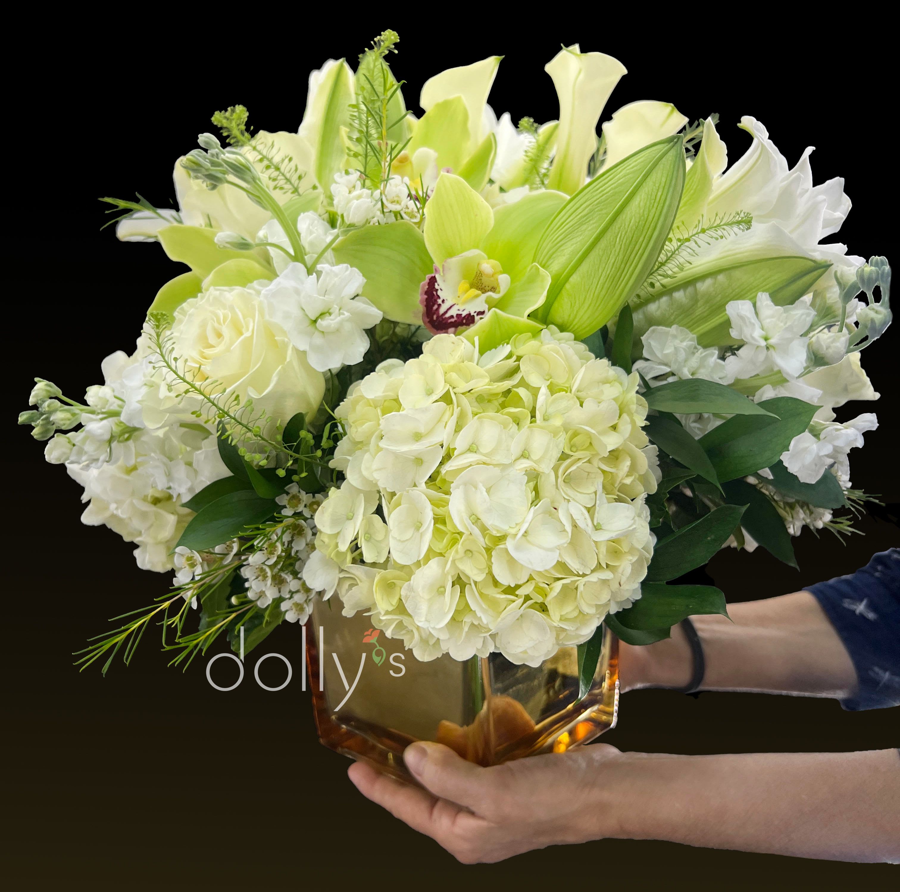 Low arrangement of white and pale green hydrangeas, roses, lilies, and an orchid in an amber glass cube vase.