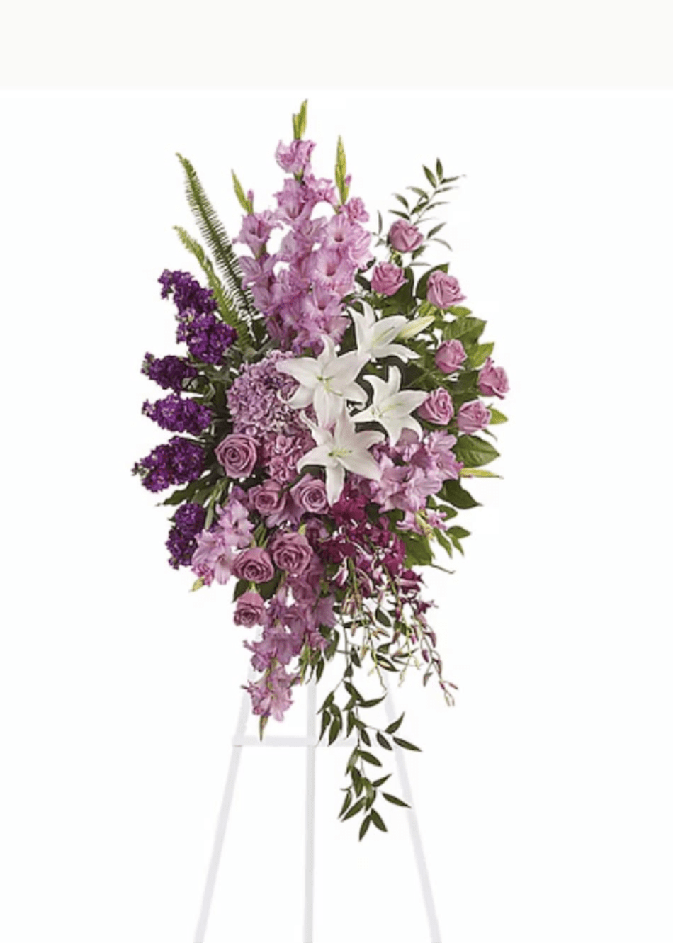 GARDEN SPRAY - Soft lavender and white blooms form a serene, formal sympathy standing spray designed to honor a loved one. This memorial spray pairs white lilies with lavender and deep purple roses, purple stock, and additional pink-purple flowers arranged in a layered, cascading silhouette on a white easel-a respectful centerpiece for a funeral service or graveside tribute.  The design is tall and vertical, dense at the center and tapering downward with trailing greenery for a graceful cascade. Colors include purple, white, pink, and green; hard goods provided are a floral stand and white easel. The arrangement's textures-from the lilies' smooth petals to the stock's ruffled clusters-create a balanced, solemn presentation that reads both elegant and comforting.  Created to give the person you're honoring a calm, dignified focal point, this spray communicates sympathy and remembrance without words. Standard care is to keep the foam hydrated until placement; please indicate delivery location and service details when ordering so we can ensure timely placement on the day of the service.