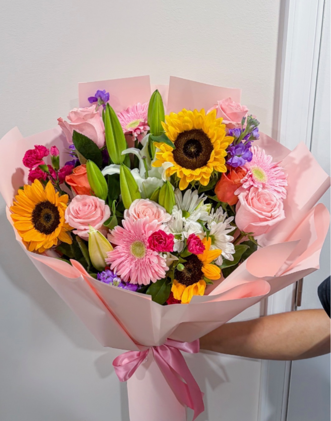 fiesta bouquet - Brilliant sunflowers set a joyful tone in this fiesta bouquet, the perfect birthday bouquet or congratulations arrangement that also reads as a spontaneous "just because" or romantic surprise. Soft pink roses and gerbera daisies bring a tender, affectionate note while pops of purple and crisp white daisies lift the composition so the person you're thinking of will feel celebrated and seen at first glance.  This hand-tied flower arrangement pairs tall unopened lily buds for height with a layered mix of sunflowers, roses, lilies, gerbera daisies, carnations, chrysanthemums and classic white daisies. The palette of warm yellow, blush pink, purple accents and white creates lively contrast and soft romance. The bouquet is wrapped in blush pink paper and finished with a pink satin ribbon for a polished, gift-ready presentation.  Ideal for birthdays, congratulations, anniversaries, or a spontaneous pick-me-up, the fiesta bouquet reads cheerful and heartfelt without being over the top. For best enjoyment, remove any packaging, recut stems, and place in fresh water; replace water and trim stems every few days to keep blooms at their brightest.