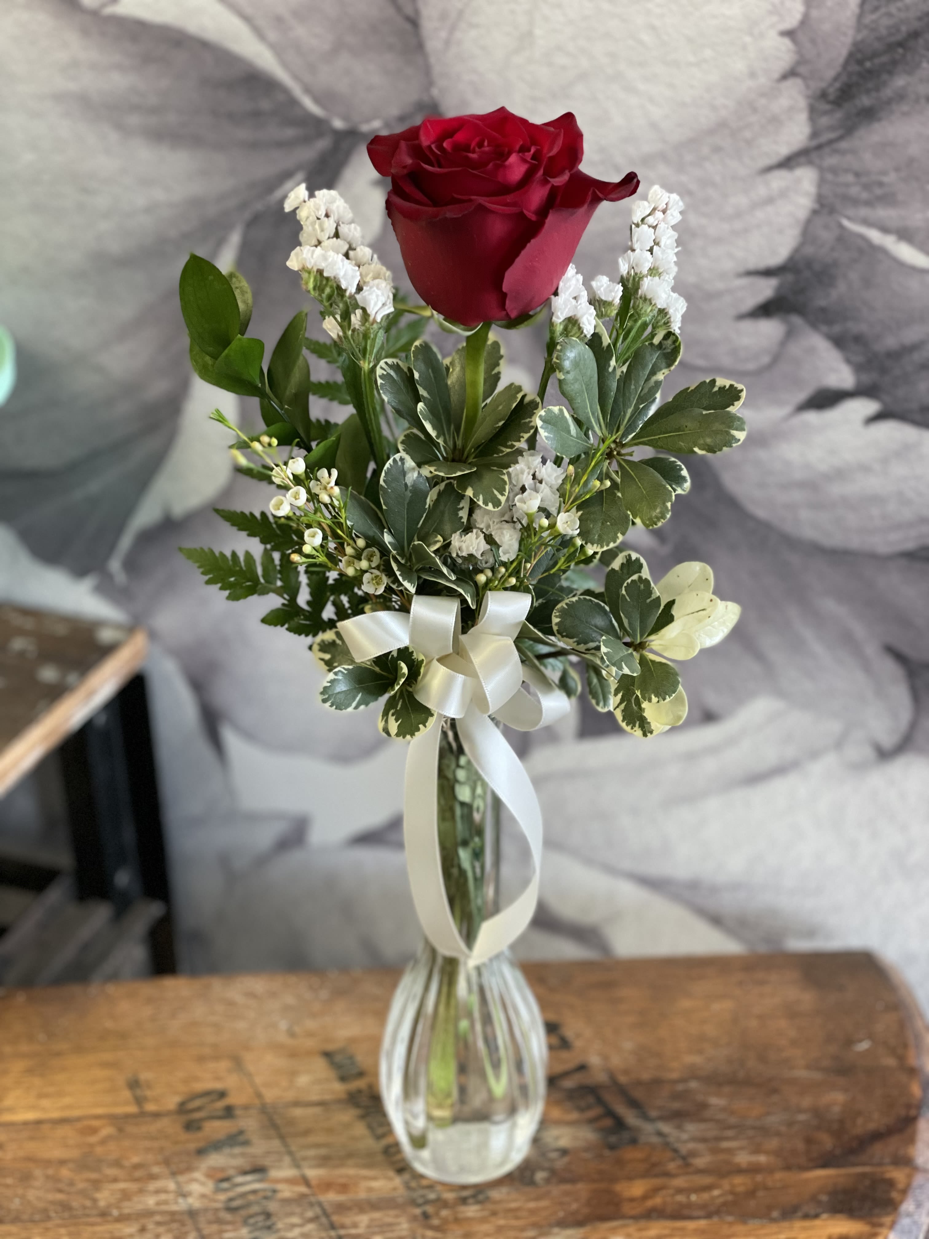 Single red rose in a clear glass vase with white ribbon and small white filler flowers