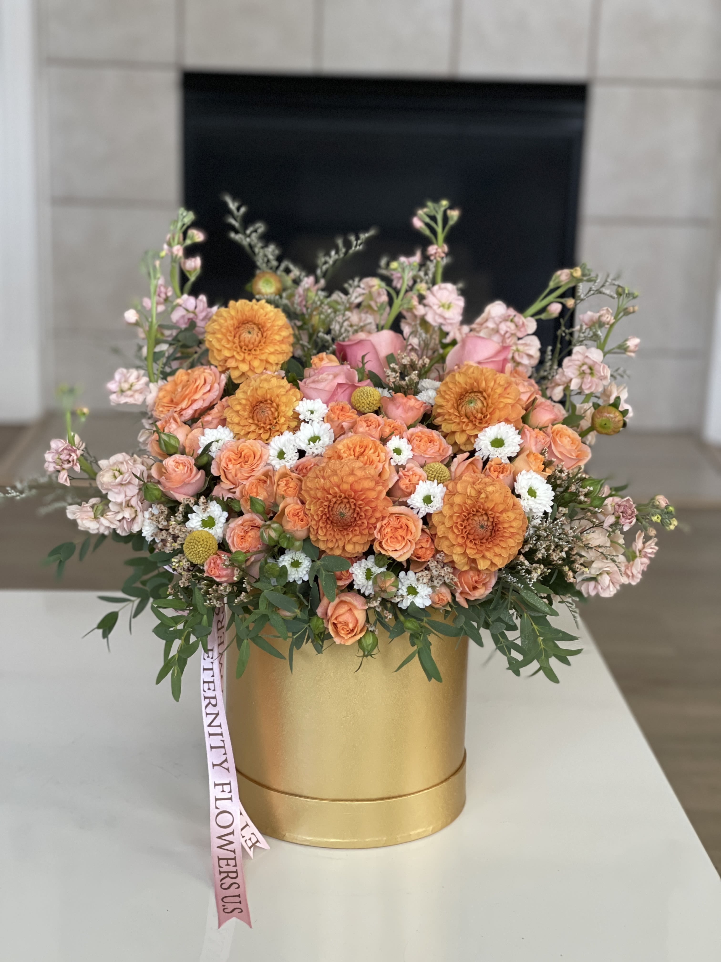 Orange and pink floral arrangement in a gold box with a ribbon