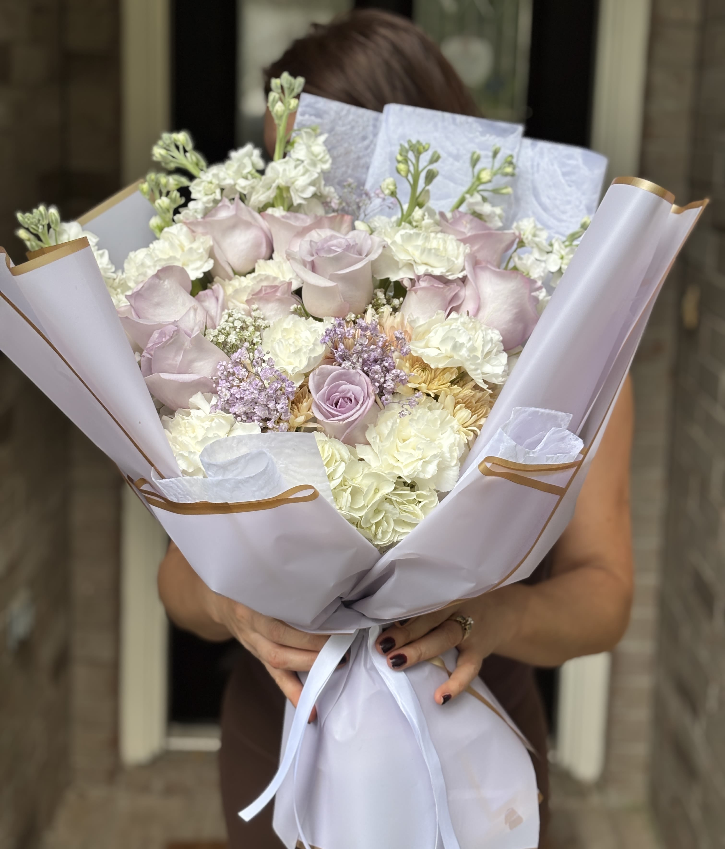 Bouquet Antonia birthday - Pale pink roses set the tone in this romantic hand-tied bouquet, a soft celebration arrangement perfect for anniversaries, springtime gestures, or simply to show sincere appreciation. The rounded, dense shape reads elegant yet effortless-the kind of gift that makes the person you're thinking of feel truly seen and cherished the moment they lift the paper.  Clusters of white carnations and tall white stock add volume and gentle vertical movement, while lavender filler and a few buttery yellow blooms introduce a whisper of contrast that keeps the palette light and fresh. Wrapped in crisp white paper with gold edging and finished with a white satin ribbon and gold-trimmed accents, the presentation is polished and gift-ready with an airy texture from the mixed blossoms.  Ideal for celebrating milestones, saying thank you, or brightening someone's day, this arrangement arrives ready to display in a vase-simply trim stems and add fresh water to extend bloom life. The compact, rounded size makes it easy to place on a bedside table, office desk, or dining table as a thoughtful, elegant surprise.