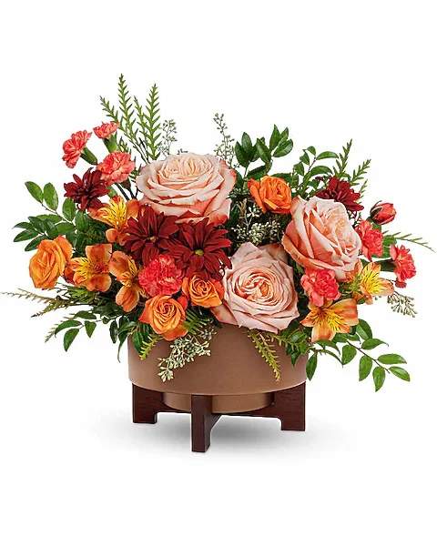 Copper Celebration - Celebrate the season with warmth and charm. The Copper Celebration Centerpiece captures fall's cozy glow with rich orange blooms and lush textures, beautifully arranged in a copper-finished planter with wooden stand-perfect for Thanksgiving gatherings and autumn moments. Copper Celebration features peach roses, orange spray roses, orange alstroemeria, miniature orange carnations, and red daisy spray chrysanthemums, it's accented with seeded eucalyptus, grevillea, and huckleberry