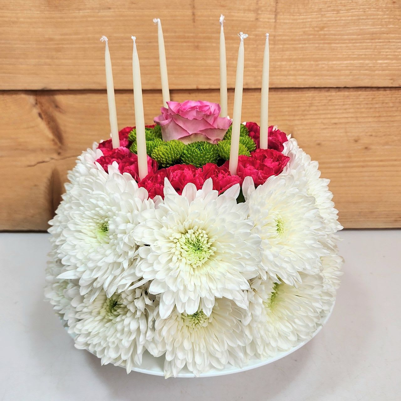 Wonderful Wishes Floral Cake - The Wonderful Wishes Flower Cake is a stunning floral arrangement crafted to resemble a beautifully decorated cake, perfect for celebrating life's special moments. Featuring a delightful blend of blooms, this elegant display showcases pristine white cremones, symbolizing purity and joy, soft pink roses, representing love and admiration, and vibrant green button mums, adding a fresh and cheerful touch. Accentuated by hot pink mini carnations, bringing a pop of lively color, this exquisite floral "cake" is artfully arranged atop a classic cake pedestal, making it a charming centerpiece for birthdays, anniversaries, or heartfelt well-wishes. A sweet and timeless gift that delivers beauty and happiness with every petal!  approx 12" H x 10" W   PLEASE NOTE: Some flowers, vases and other products pictured are subject to availability, and may be substituted for a similar item if unavailable according to our substitution policy.