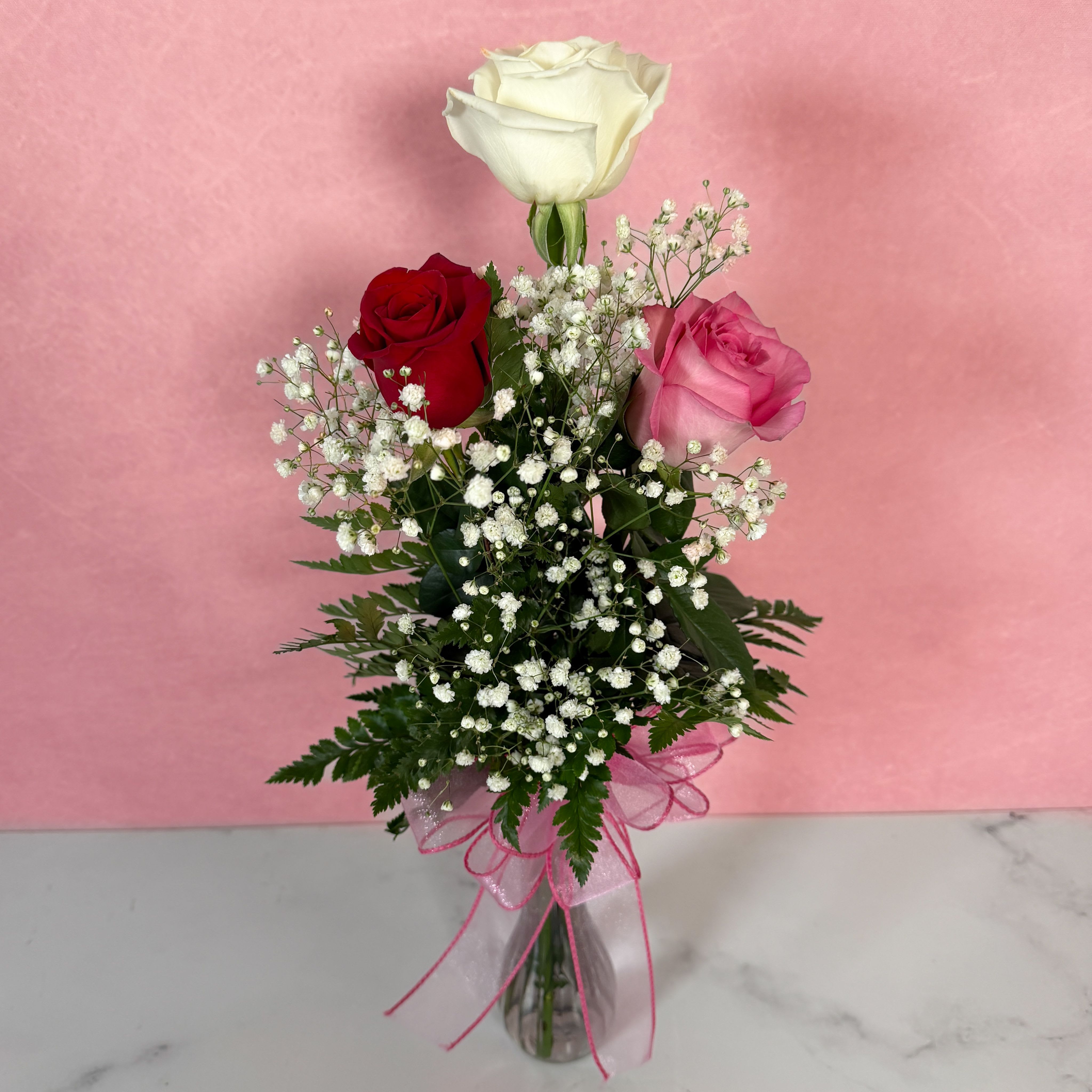 Three roses in a clear vase with baby's breath and a pink ribbon