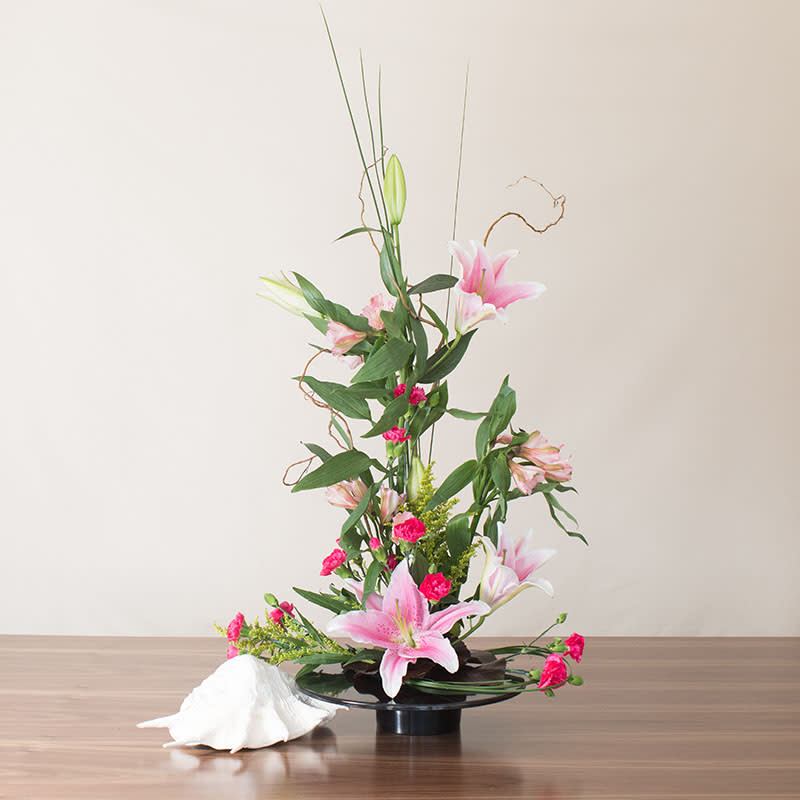 Ikebana-style arrangement of pink lilies and carnations in a low black dish with a white seashell accent.