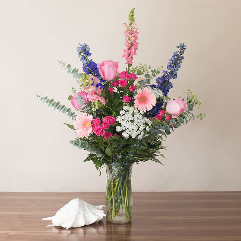 Tall pink, blue, and white mixed flower arrangement in a clear glass vase on a table