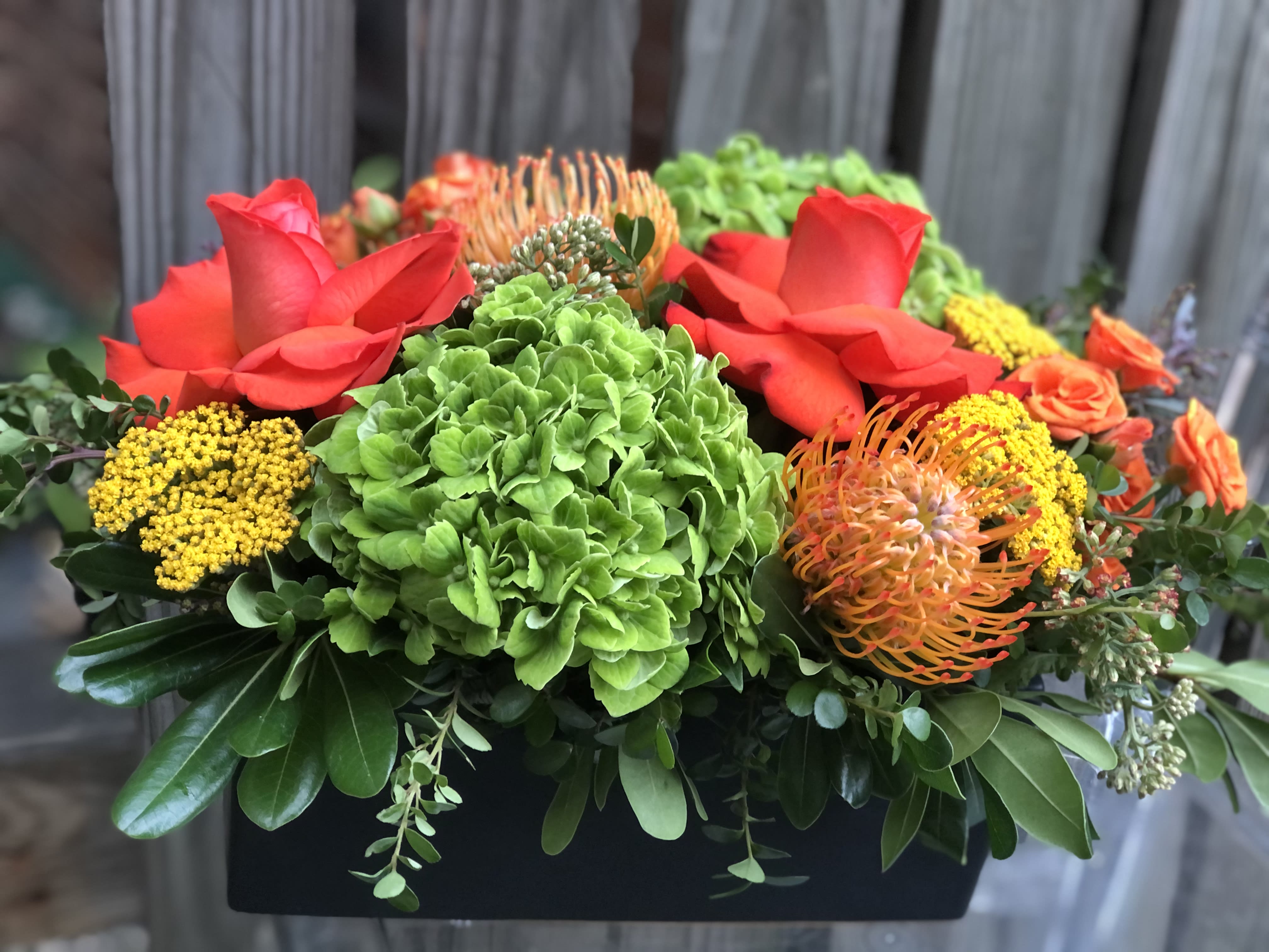 Bright mixed floral arrangement with red roses, green hydrangea, and orange pincushion blooms in a black container.