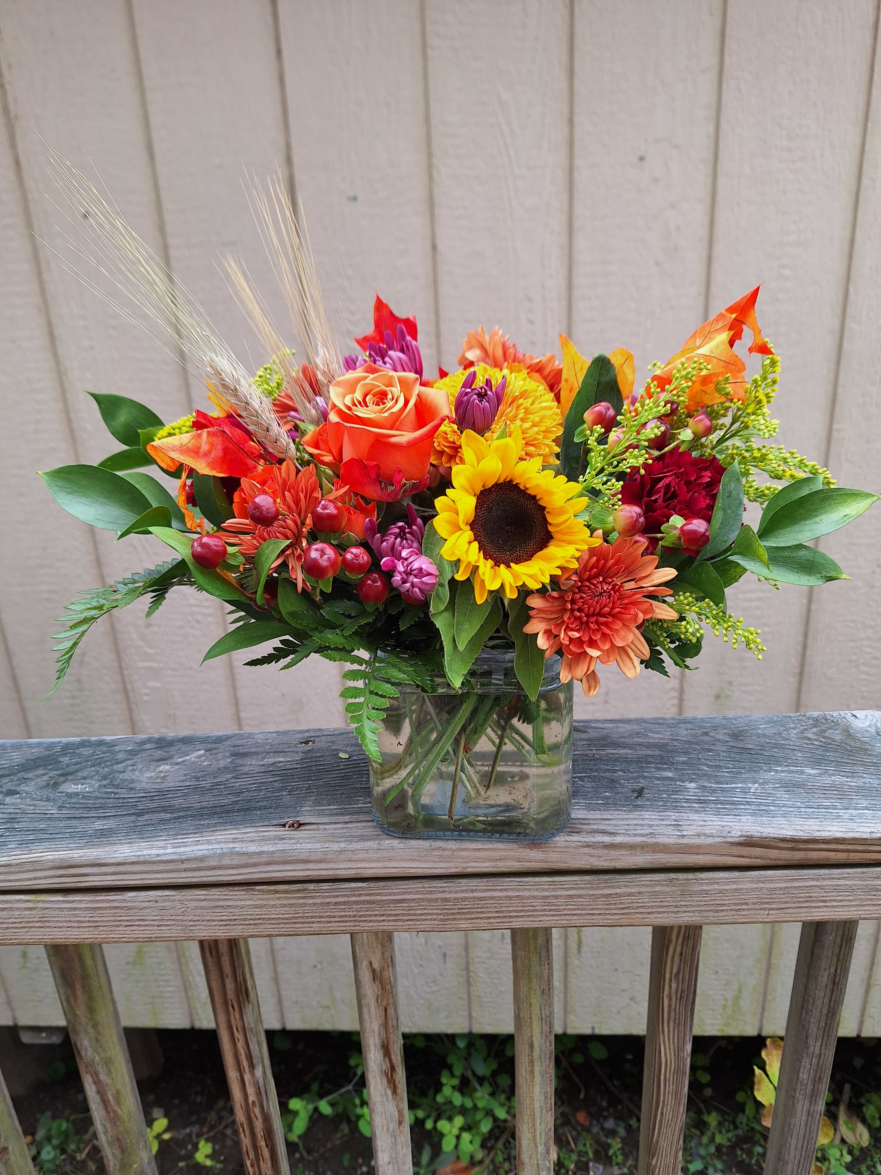Bountiful Fall Cube Arrangement - Brilliant yellow sunflowers, orange roses, gold and bronze chrysanthemums, burgandy carnations, lavender cushions, red hypericum berries, solidago, faux autumn leaves and sprigs of wheat arranged in a cube glass vase.