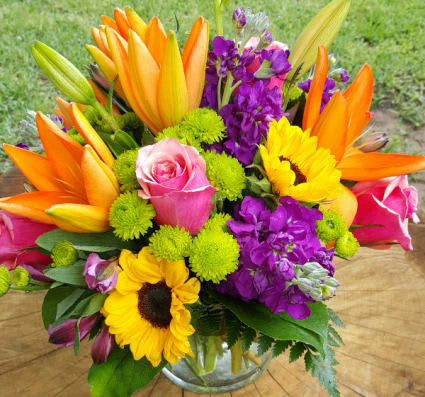 Bright mixed bouquet with orange lilies, pink roses, purple flowers, and sunflowers in a glass vase
