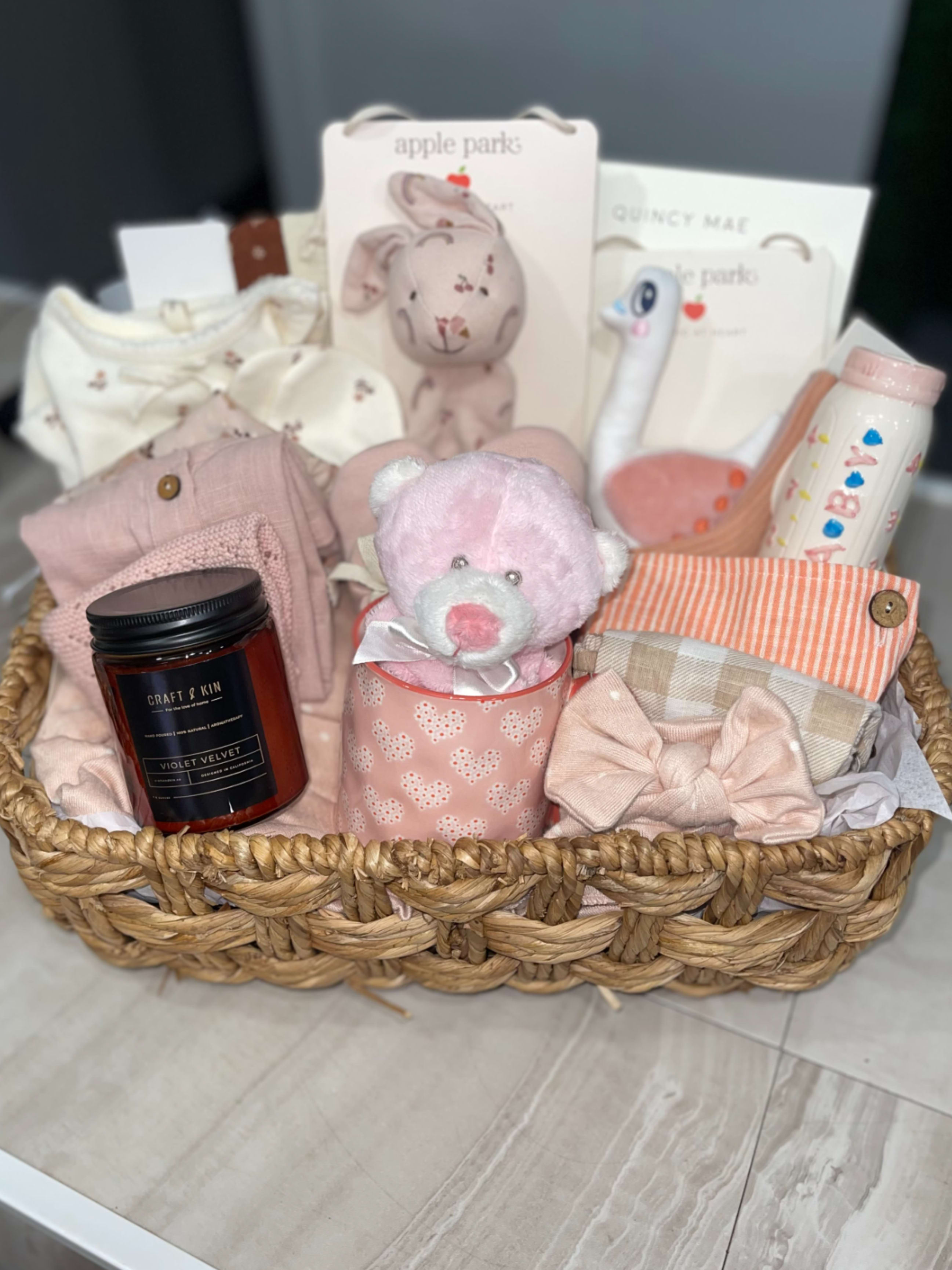 Large Hello Baby Girl Basket - Soft pastels and cuddly textures make this Hello Baby Girl Basket the kind of newborn gift that instantly comforts and delights. Perfect as a new baby or baby shower present, the baby gift basket reads like a thoughtful welcome-home surprise for the little girl you're celebrating - she'll be surrounded by soft blankets, plush friends, and sweet essentials that set a gentle, nursery-ready tone.  Arranged upright in a white wire or woven basket, this set includes a plush teddy or doll, folded baby clothing and blankets in pink, mint, lavender, peach and cream, plus curated extras like a scented candle jar, packaged accessories. The layered presentation and mixed textures - soft knits, cotton garments, and wicker basket - create a tidy, gift-ready look that's easy to place in the nursery. And A variety of mixed blooms and foliage in a glass bud vase.  This basket is ideal for the new mom who could use cozy essentials and a little pampering; it's the kind of present that says "congratulations" while helping baby feel snug and loved. 