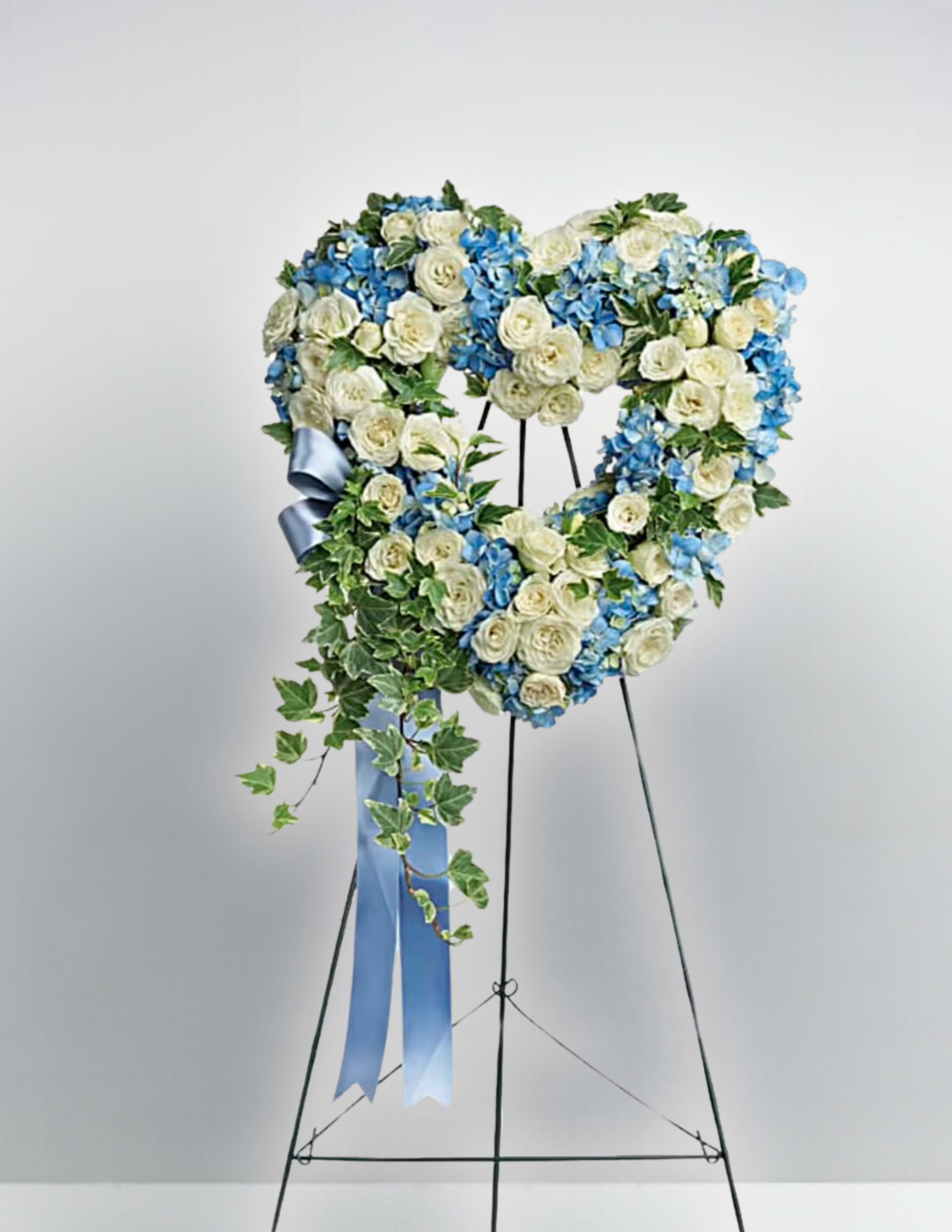 Heart-shaped floral wreath of white and blue flowers on a stand with a blue ribbon