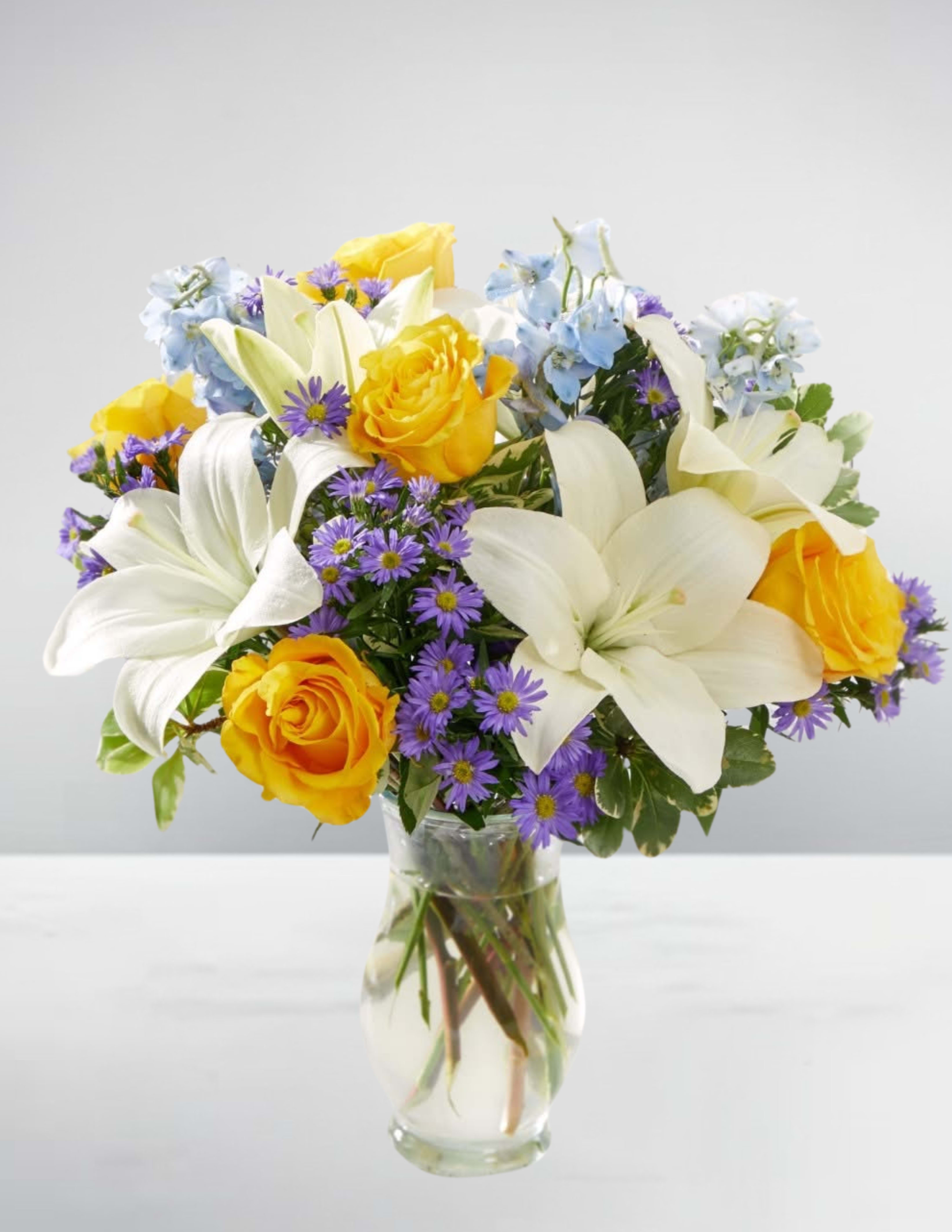 Sunny delight - The bouquet, named "Sunny Delight," is a vibrant and cheerful arrangement that exudes warmth and happiness. The clear glass vase is filled with a colorful mix of flowers, including bright yellow roses, elegant white lilies, and delicate purple asters. The stems of the flowers are visible through the vase, adding a touch of natural beauty to the overall design.  The bouquet is placed on a white surface, which provides a clean and neutral background that allows the flowers to take center stage. The plain white wall behind the bouquet adds to the simplicity and elegance of the overall presentation.  The "Sunny Delight" bouquet is perfect for brightening up any room or occasion. Its vibrant colors and delicate textures create a sense of joy and happiness, making it an ideal choice for gifts, decorations, or simply adding a touch of warmth to your home.