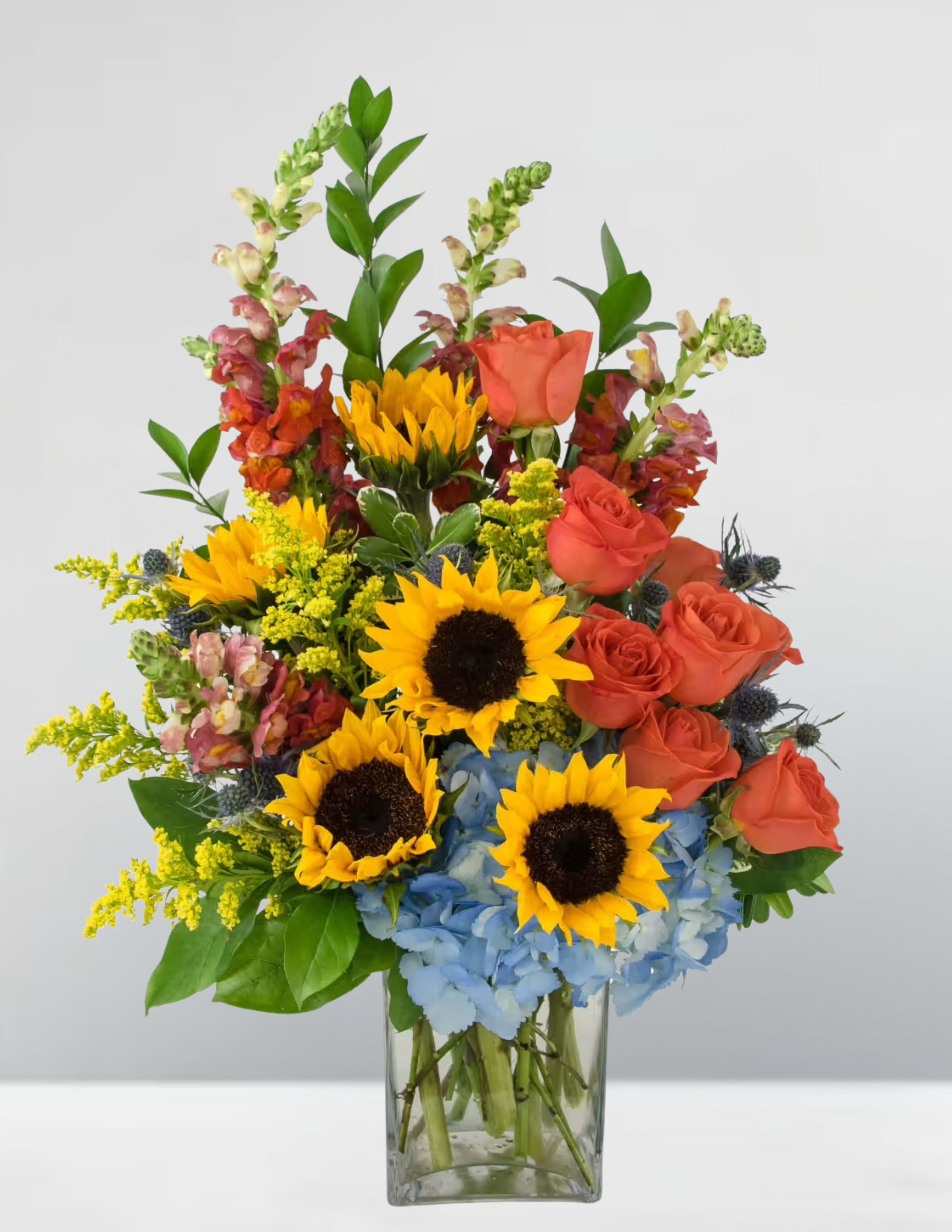Sunflowers, orange roses, and blue hydrangeas in a clear glass vase