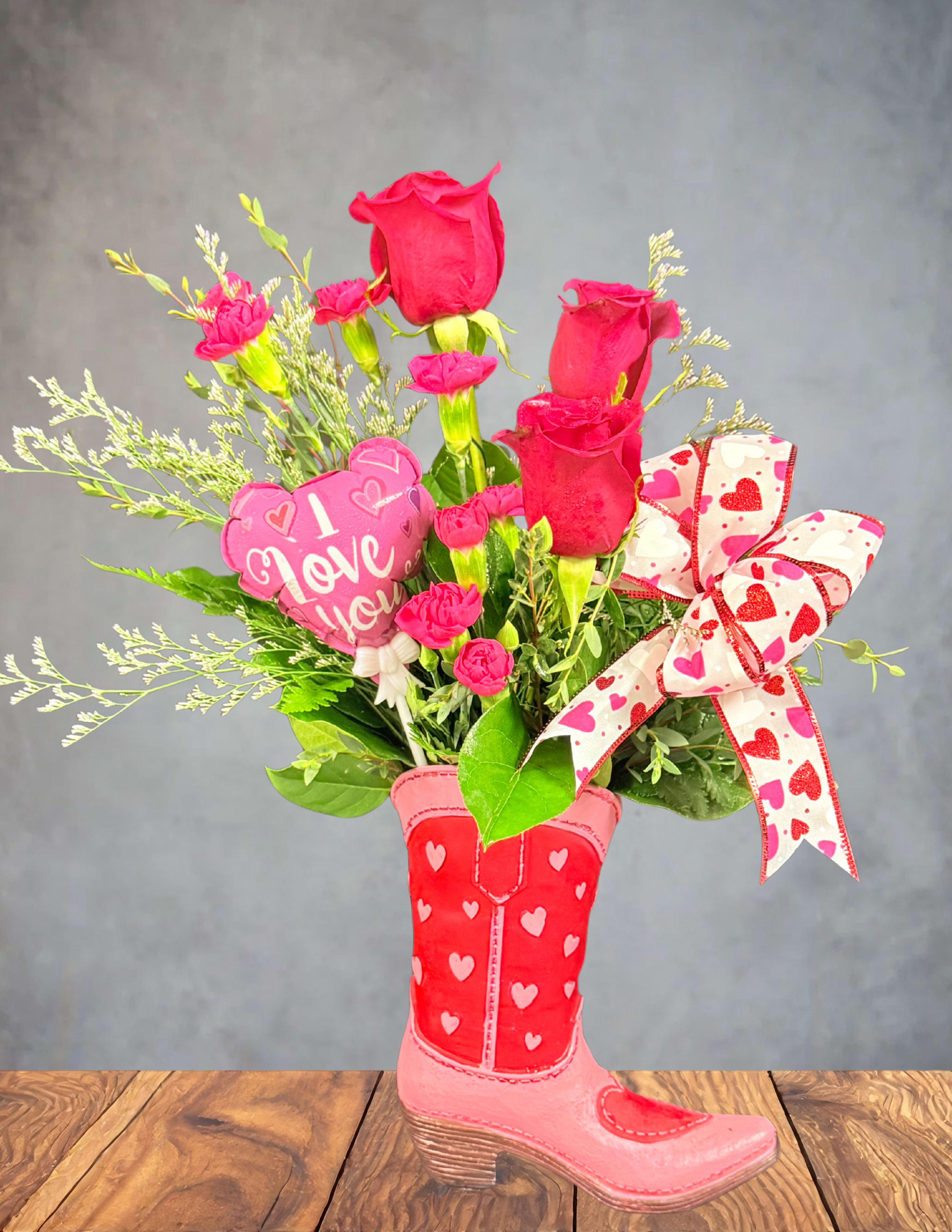 Pink roses and carnations arranged in a heart-decorated boot vase with a ribbon.