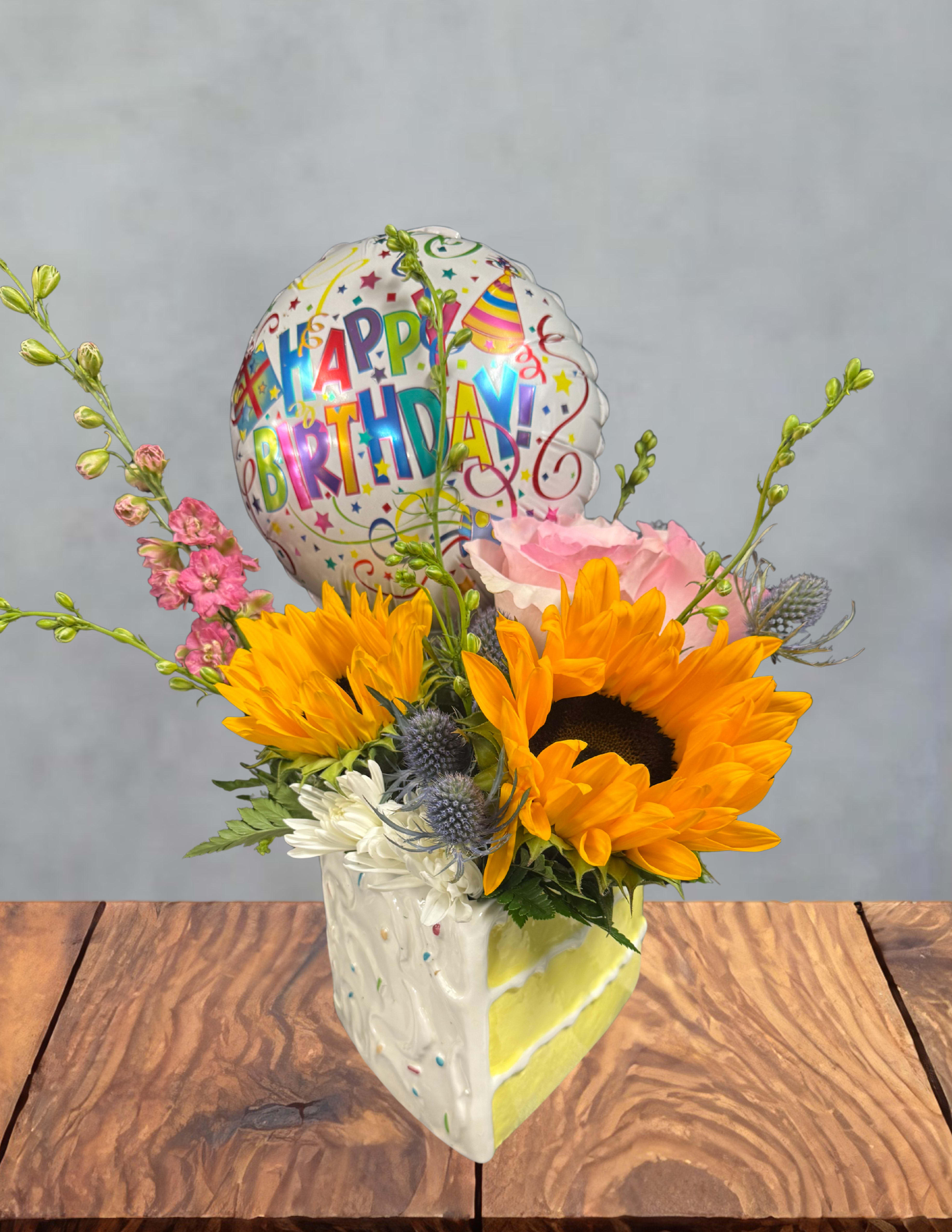 Confetti surprise  - A burst of sunshine and party cheer-this birthday bouquet pairs bold sunflowers with white daisy-like blooms, a single pink rose, and delicate purple accents to give the recipient an instant lift. As a birthday arrangement, Confetti Surprise reads as joyful and celebratory: warm yellows and soft pinks mingle with cool purples so she feels seen and celebrated on her special day.  The flowers arrive arranged in a hand-painted ceramic vase that adds playful, colorful charm to any table, plus a round "Happy Birthday" foil balloon that rises above the stems for extra delight. Textures range from the sunflower's sturdy faces to the daisy's light petals and the rose's classic softness; colors include bright yellow, pink, white, and purple for a lively, layered look.  Perfect for a friend, family member, or colleague whose birthday you want to make memorable, this arrangement makes a cheerful centerpiece and is ready to display on arrival. Vase dimensions and care: remove balloon and trim stems every few days, refresh water and floral food as needed to extend freshness.
