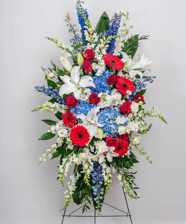 Love and honor standing spray - A tall, fan‑shaped standing spray that commands attention with a patriotic palette-perfect as a sympathy or memorial flower arrangement. The vertical composition of red roses and red gerbera daisies against crisp white lilies and clusters of saturated blue hydrangea and delphinium creates a formal, respectful statement for a funeral service, memorial, or ceremonial tribute that honors and comforts the person you're remembering.  This arrangement is densely layered for high impact: glossy broad foliage and trailing white filler give fullness and soften the structure while keeping the dramatic color contrast intact. The mix of roses, gerbera daisies, lilies, hydrangeas and delphinium provides varied texture-from the rounded hydrangea clusters to the sculptural lilies and delicate delphinium spikes-mounted on a sturdy metal easel for display.  Choose this spray when you want the recipient or congregation to feel recognized and respected-a meaningful way to express love and honor at a service. Standard sizing suits most display needs; pick delivery timed for the ceremony and follow basic care: keep flowers sheltered from extreme heat and place on the easel at the venue for best presentation.