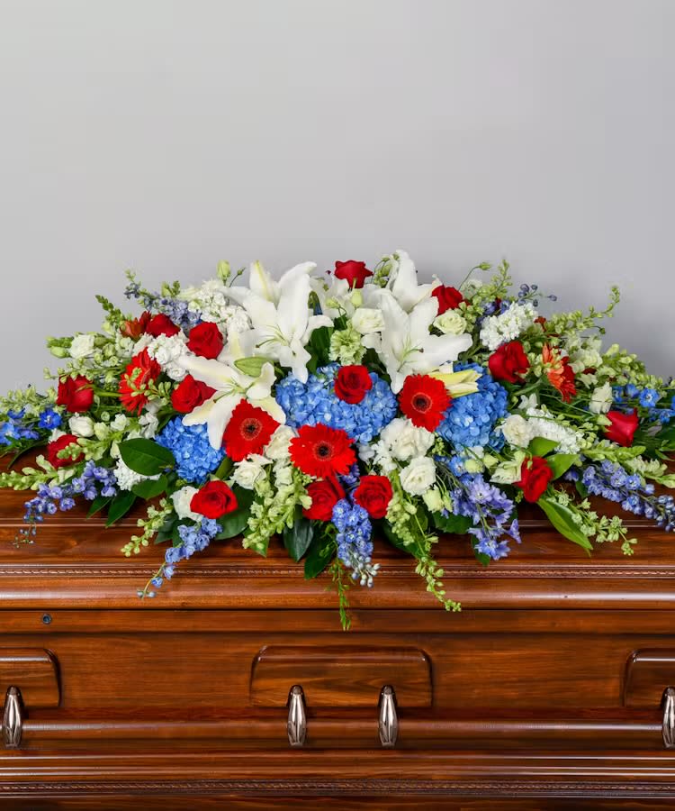 Love and honor casket spray - A broad, low-profile casket spray that reads as a dignified salute-white lilies and red roses take center stage in this funeral arrangement, making it an appropriate choice for a memorial or funeral service. The red, white, and blue palette conveys love and honor while the full, formal silhouette drapes across the polished wooden casket to create a respectful, ceremonial presence the person you're remembering will receive with quiet reverence.  This flower arrangement combines trumpet-shaped white lilies, classic red roses, vivid blue hydrangeas, red gerbera daisies, and supportive carnations for texture and volume, all arranged in a lush, cascading composition. Mixed greenery frames the blooms and softens the edges, giving the spray a balanced, layered look that reads well from the front of the service. The design is substantial in scale to cover a casket top and is fashioned for a polished, formal presentation.  Made specifically for funeral and memorial settings, this casket spray offers a meaningful way to express sympathy and respect-ideal from family, close friends, or organizations wanting to honor a life. Care and handling are managed by our florists to keep blooms fresh through the service; please provide delivery details and service time when ordering so we can ensure timely placement.