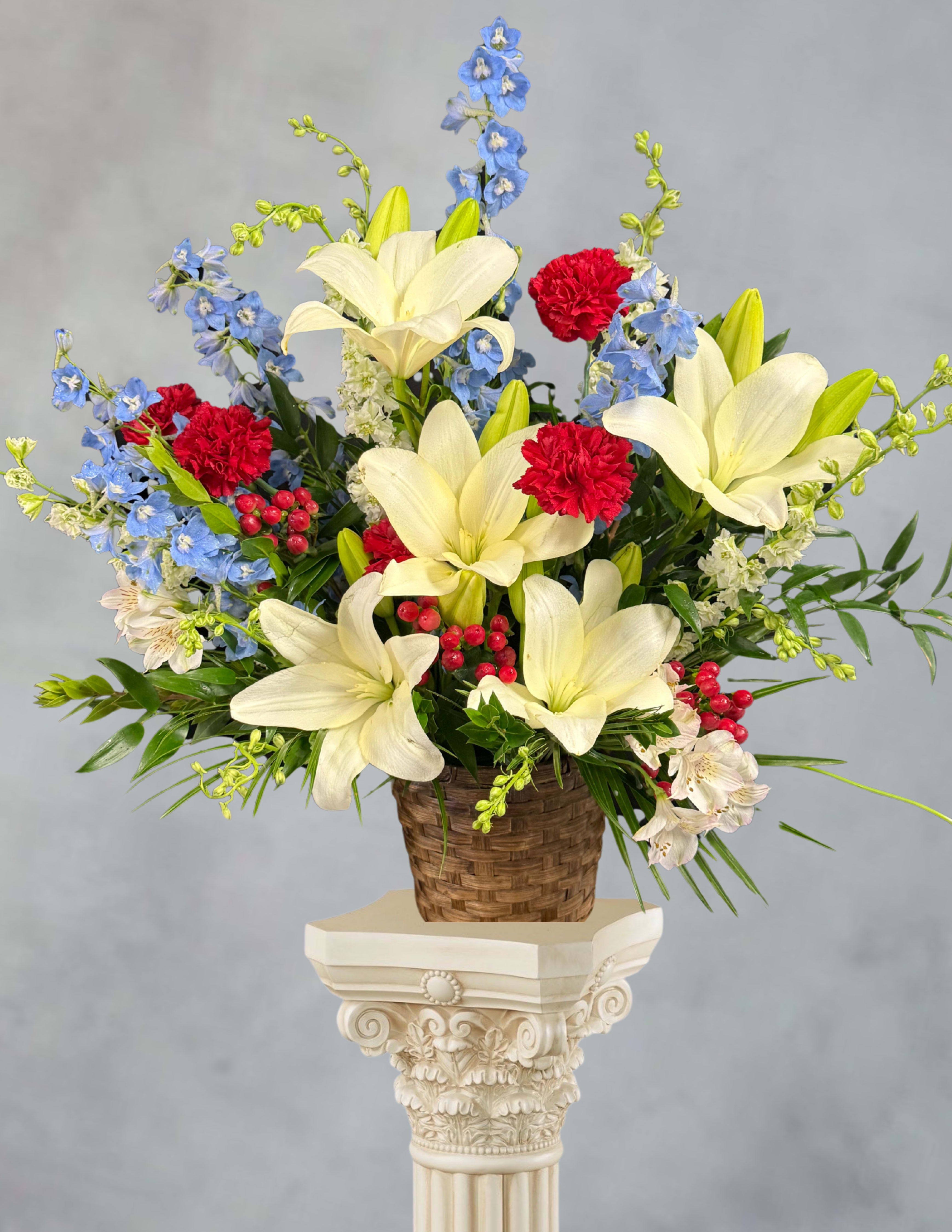 Draped in honor - Crisp white lilies and towering blue delphinium spikes create a striking red-white-blue statement perfect for a patriotic tribute, congratulations display, or a proud anniversary bouquet. The upright, broad composition reads formal and celebratory-the kind of flower arrangement that makes the person you're honoring feel seen and applauded. Small red berry clusters and airy white filler weave through the design to add movement and texture, so the recipient experiences both bold color and delicate detail.  This hand-arranged piece sits in a woven basket and can be showcased on the included decorative pedestal stand for added presence; its natural basket base keeps the look grounded and tasteful. Alongside the lilies and delphinium you'll find bright red carnations and complementary greenery that give fullness and lasting visual impact. The balanced spread extends outward for an elegant silhouette that works well on entry tables, stage displays, or as a centerpiece at a memorial or celebration.  Choose this arrangement when you want the recipient to feel honored-whether for a Fourth of July tribute, a promotion, or to mark a meaningful life moment. As a fresh-flower composition, stems benefit from fresh water and a cool room away from direct sunlight to help blooms open fully and last longer; please indicate any delivery notes at checkout for placement on a pedestal stand.