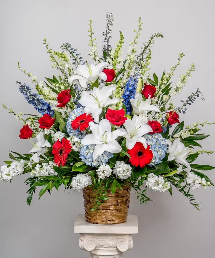 Love and honor sympathy basket - A tall, formal sympathy arrangement in a woven basket and on a metal easel, this Love and honor sympathy basket presents classic red roses, bright red gerbera daisies and pure white lilies layered with blue hydrangea clusters and sky‑blue delphinium spikes. The red, white and blue palette reads as both ceremonial and comforting-perfect for a memorial service, patriotic tribute, or to honor someone who cherished bold, traditional florals. The vertical, full composition and broad dark foliage create a dignified silhouette that will make the recipient feel deeply remembered and respected.  Rich texture comes from lush hydrangeas and airy delphinium above a foundation of roses, lilies, stock and mixed filler flowers, all artfully arranged with abundant greenery in a substantial woven basket and supported on a sturdy metal stand or floral easel for upright display. This large-scale piece is designed to be seen from all sides and holds its shape well-ideal for a funeral program, graveside service, or to stand beside a home altar. Standard care: keep in a cool place away from direct sun and replenish water as needed for the cut stems to prolong freshness.