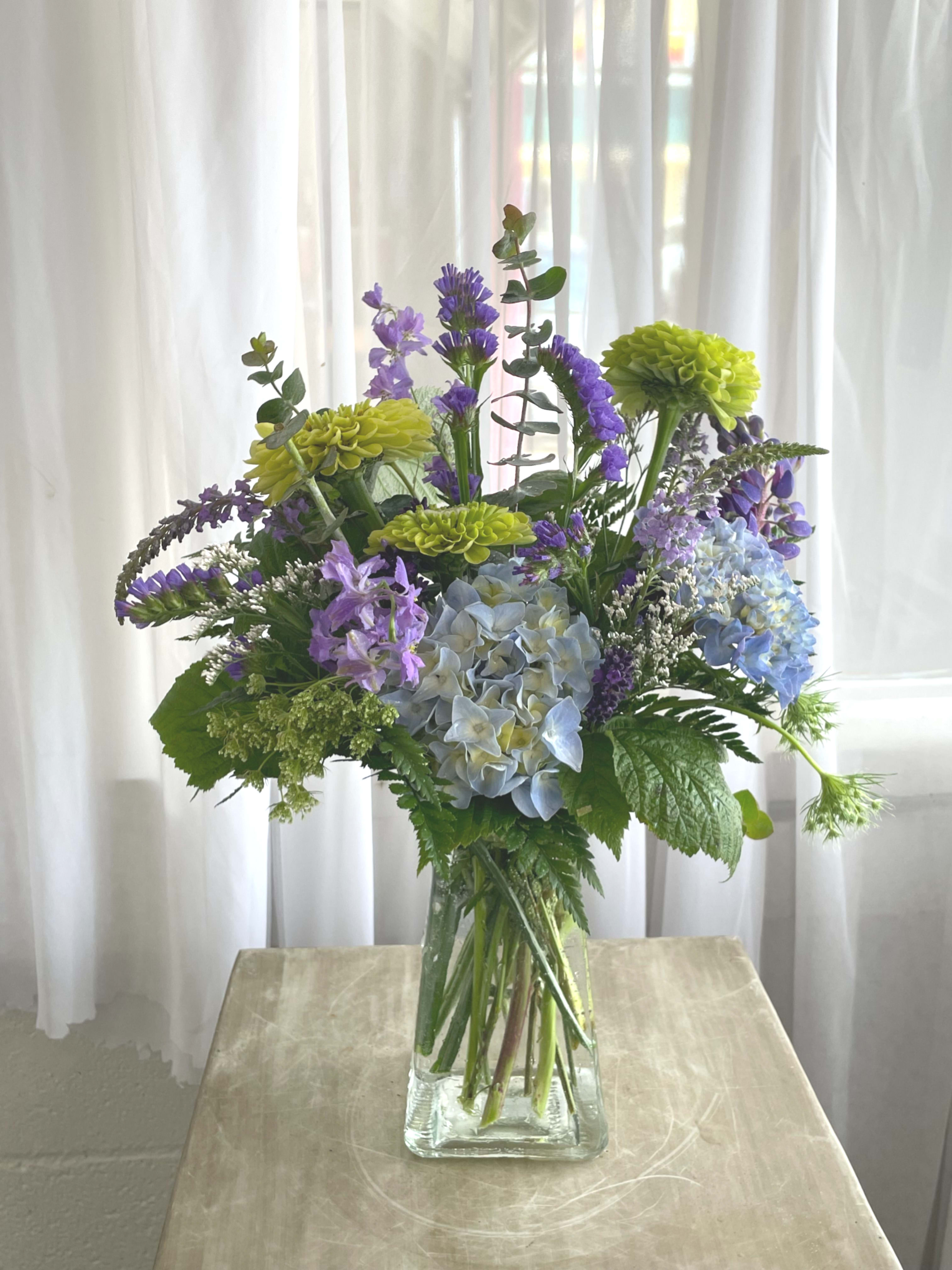 Mixed bouquet of blue hydrangeas, purple flowers, and green blooms in a glass vase