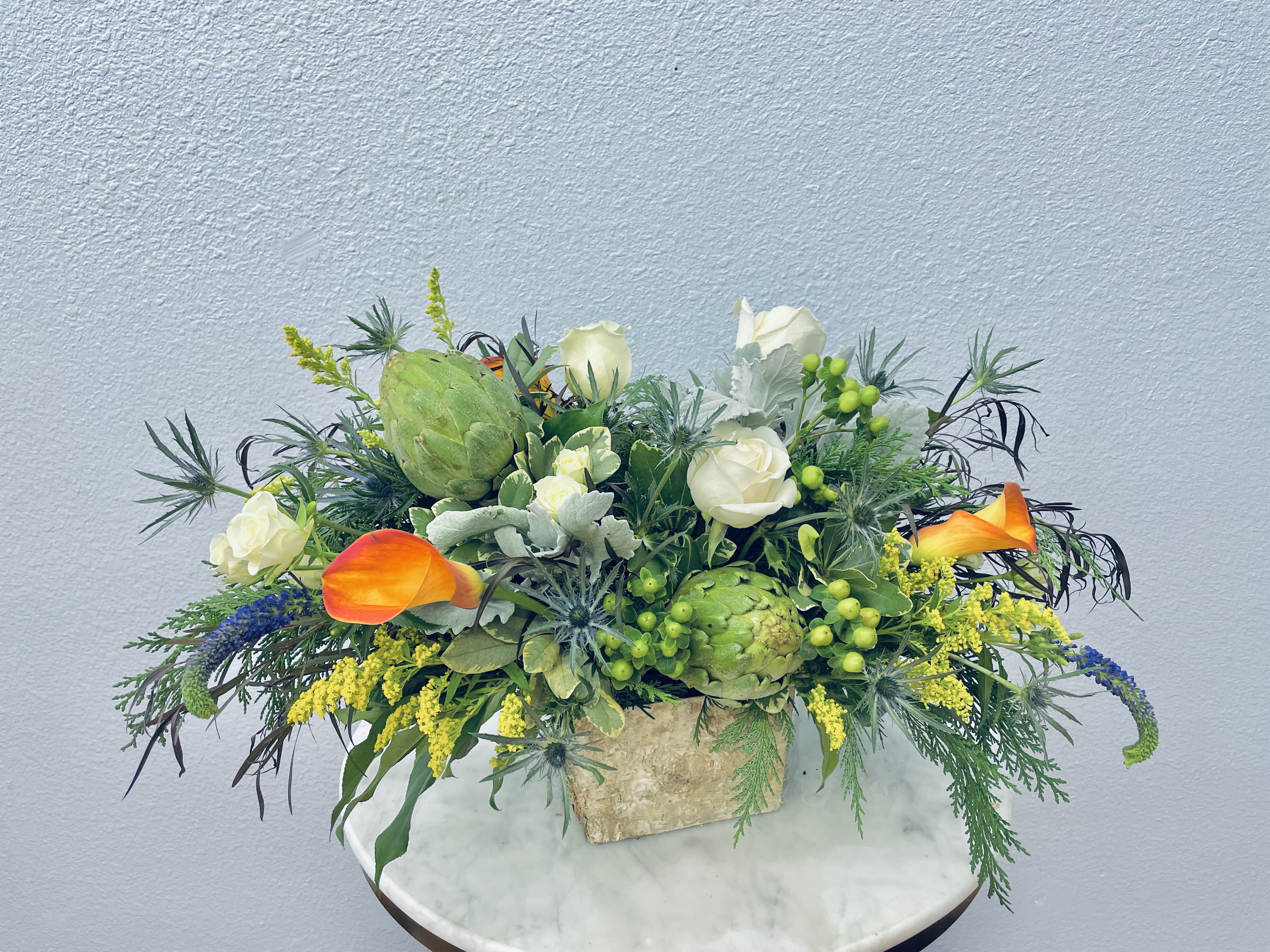 Mixed floral arrangement with white roses, orange calla lilies, and green artichokes in a stone vase