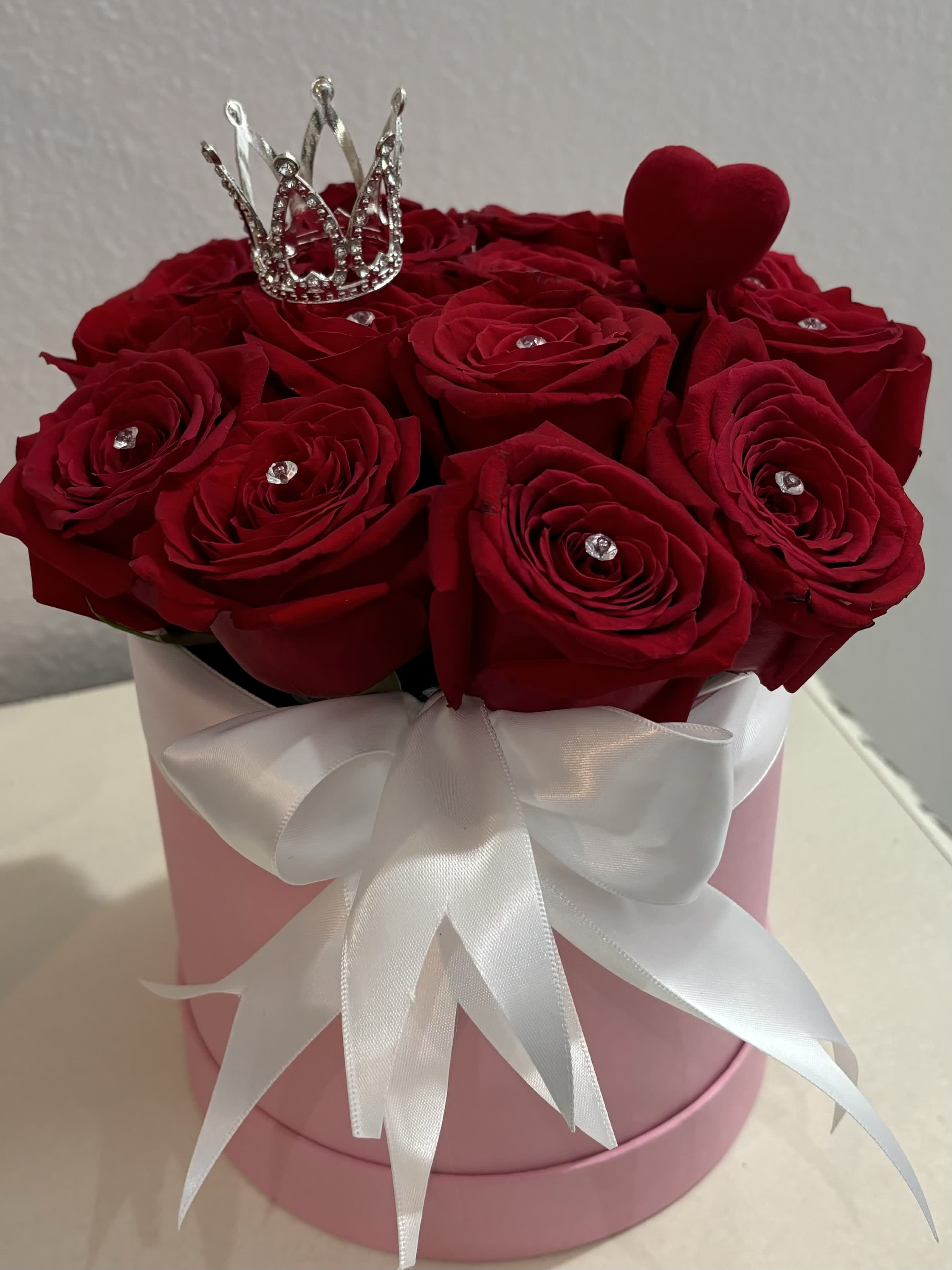 Red roses in a pink hat box with a white ribbon, crown, and heart topper