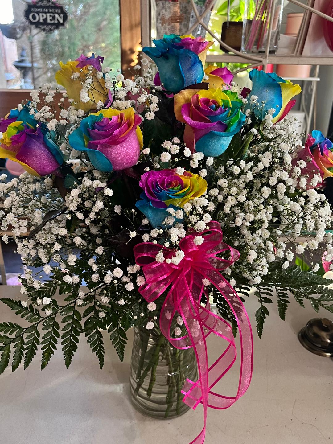 Rainbow-colored roses in a glass vase with baby's breath and a pink ribbon