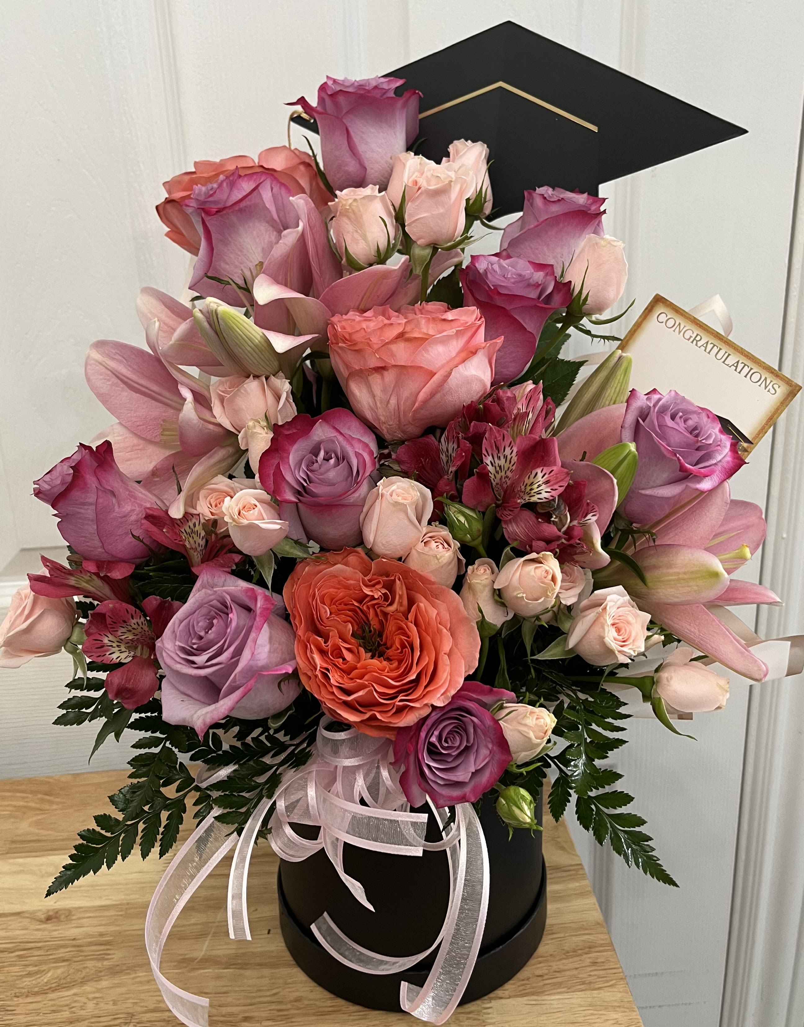 Pink and lavender roses with lilies in a black hatbox, topped with a graduation cap.