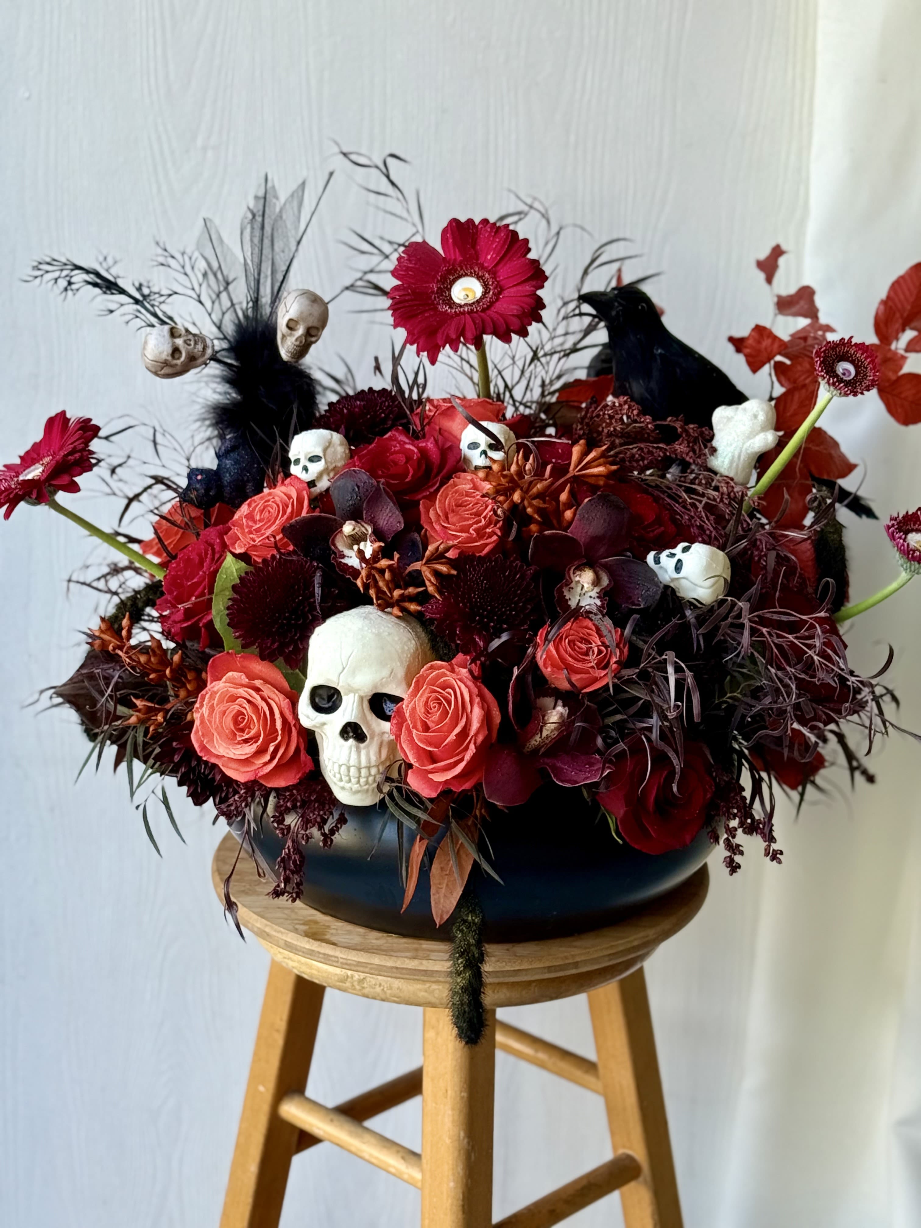Dark Enchantment Halloween Floral Arrangement - Make a bold statement this Halloween with our Dark Enchantment floral arrangement — a large, luxurious centerpiece bursting with orange roses, red gerbera daisies, cymbidium orchids, and strawflowers, all surrounded by dark, moody textures. Designed in a sleek black container, it’s accented with skulls, raven, and eerie details for the perfect mix of elegance and fright.This large arrangement overflows with orange roses, red gerbera daisies, deep-toned blooms, and cymbidium orchids, surrounded by eerie textures and dark foliage. this piece perfectly blends luxury and spookiness.  Ideal for Halloween parties, haunted house décor, or dramatic seasonal displays, this arrangement adds instant atmosphere and sophistication.