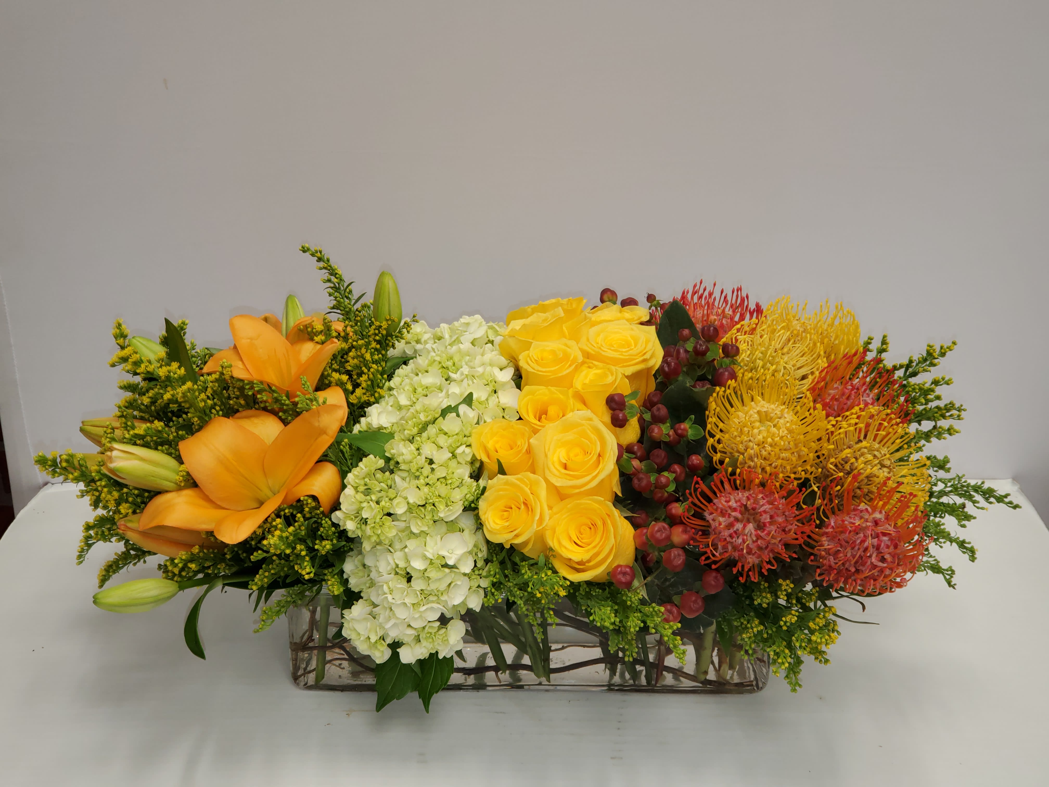 Long floral centerpiece with orange lilies, yellow roses, and mixed blooms in a rectangular vase