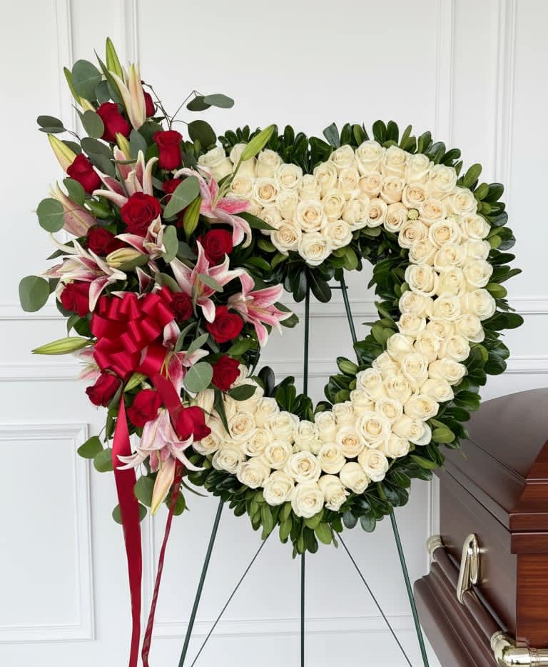 Eternal Devotion Heart - A stunning expression of everlasting love, this luxurious heart-shaped wreath is crafted with an abundant display of soft white roses, symbolizing purity, honor, and eternal remembrance. Accented with a dramatic cluster of rich red roses and elegant lilies, this design beautifully represents deep love, respect, and devotion.  Lush greenery and flowing ribbon details add depth and movement, creating a striking and sophisticated tribute. The heart shape serves as a powerful symbol of an unbreakable bond, making this piece a meaningful way to honor a life that will always be remembered.  Available in multiple sizes, this arrangement offers a refined and heartfelt expression of sympathy, love, and enduring connection.