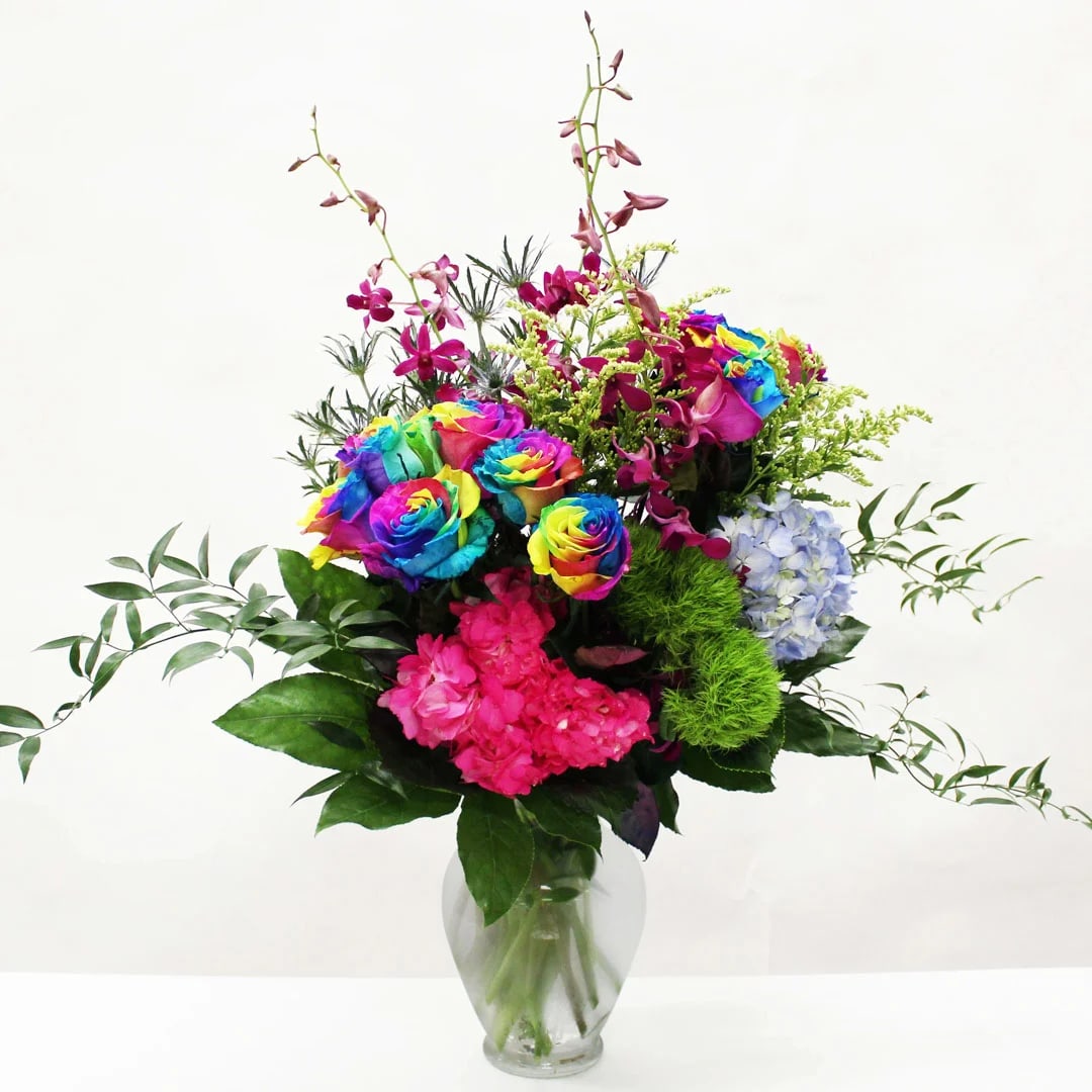 Tall arrangement of rainbow roses, orchids, and bright mixed blooms in a clear glass vase