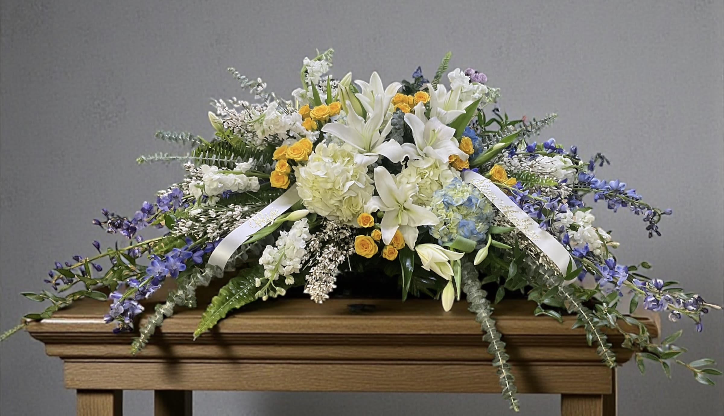 Angelic Embrace Casket Spray - Angelic Embrace is a soft, refined casket spray designed with a calming blend of whites and gentle blues, creating a peaceful, balanced presence for a final farewell. Layered blooms and natural movement give the design a light, flowing feel that rests beautifully across the casket without feeling heavy or overdone.  The cool palette brings a sense of quiet comfort and serenity, offering a graceful expression of love, remembrance, and respect. Thoughtfully arranged for proportion and placement, this piece creates a focal point that feels composed, intentional, and deeply appropriate for the setting.  A meaningful choice for honoring a life with dignity, Angelic Embrace is designed to bring calm, beauty, and quiet reassurance to the service.  Available for local delivery through Aspen Tower Florist, serving Greensburg and Shelbyville along with Rushville, Batesville, Westport, Oldenburg, St. Paul, Waldron, and Milroy.