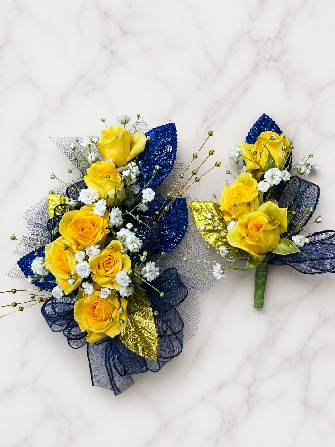 Royal Legacy Formal Set - MAGNETIC BOUTONNIERE WARNING: CONTAINS STRONG MAGNETS. DO NOT USE IF YOU HAVE A PACEMAKER, DEFIBRILLATOR, INSULIN PUMP, COCHLEAR IMPLANT, NEUROSTIMULATOR, OR OTHER IMPLANTED MEDICAL DEVICE. KEEP AWAY FROM ELECTRONICS, CREDIT CARDS, HOTEL KEY CARDS, AND CHILDREN.  Royal Legacy  Sunny yellow roses, rich navy ribbon, gold accents, and delicate filler flowers come together in Royal Legacy, a bold corsage and boutonniere collection for prom, homecoming, dances, banquets, weddings, and formal events.  Standard includes a wrist corsage with 3 spray roses and a matching magnetic boutonniere with 2 roses. Deluxe includes 4 spray roses in the corsage. Premium includes 5 spray roses for additional fullness and flower coverage. All boutonnieres include 2 roses.  Because each corsage and boutonniere is individually designed, no two sets will be exactly alike. Ribbon, filler flowers, bracelets, decorative accents, and other design elements may vary based on availability. While individual details may differ, the overall color palette, roses and/or flowers, style, and appearance of the Royal Legacy collection will be maintained.  NOTE: If spray roses arrive unusually small, additional blooms may be used to maintain the intended size and fullness of the design.