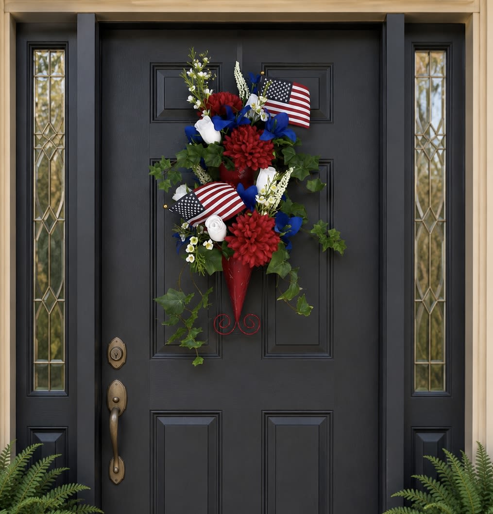 American Spirit - American Spirit isn’t for people who decorate because the calendar says it’s time. It’s for people who still stand when the flag passes by. The people who tear up during the National Anthem, wave from the parade route, and believe that home should reflect what matters most.  Designed in a bold red metal double-cone wall pocket, this striking silk arrangement features vibrant red, white, and blue florals, flowing greenery, and American flags that create an unforgettable first impression. Measuring approximately 28 inches long and 15 inches wide, American Spirit commands attention from the street while offering a warm welcome at the door.  This is more than seasonal décor. It’s a conversation piece. A statement of pride. A daily reminder of freedom, sacrifice, family, and faith in the values that built this nation. The dramatic vertical design offers a fresh alternative to the ordinary wreath while delivering year-after-year beauty without watering, fading, or maintenance.  Whether displayed for Memorial Day, Independence Day, Veterans Day, military homecomings, or throughout the year, American Spirit transforms an ordinary entrance into something meaningful. Guests won’t simply notice it—they’ll remember it.