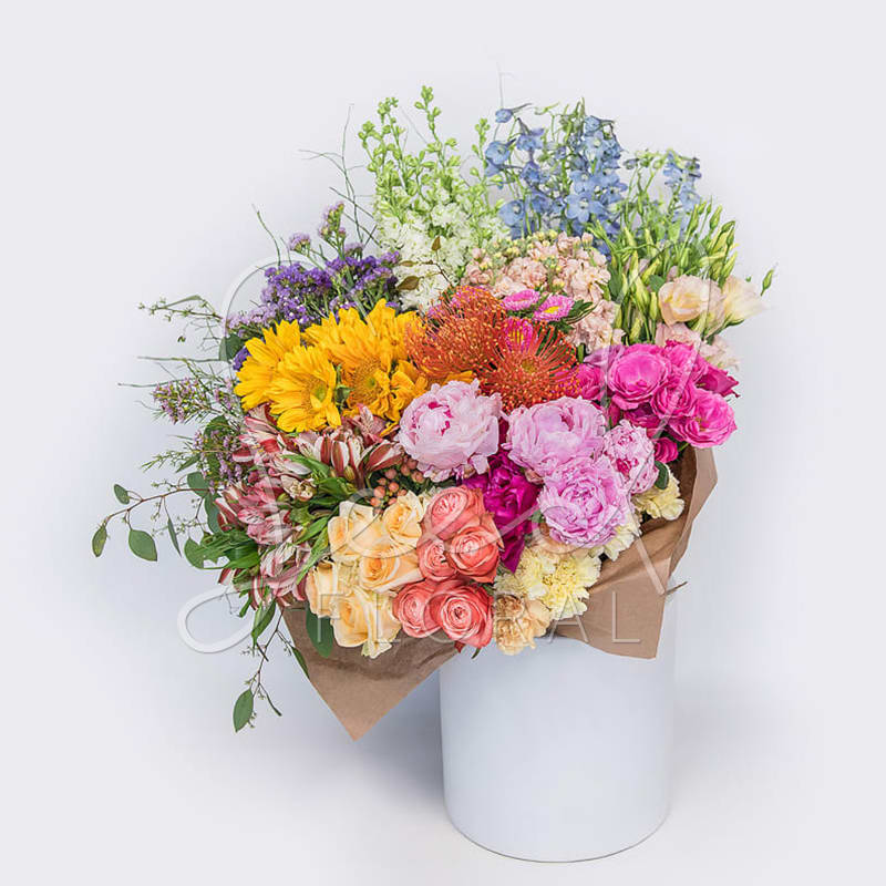 Large mixed-color bouquet with sunflowers, roses, peonies, and protea wrapped in kraft paper in a white cylinder.