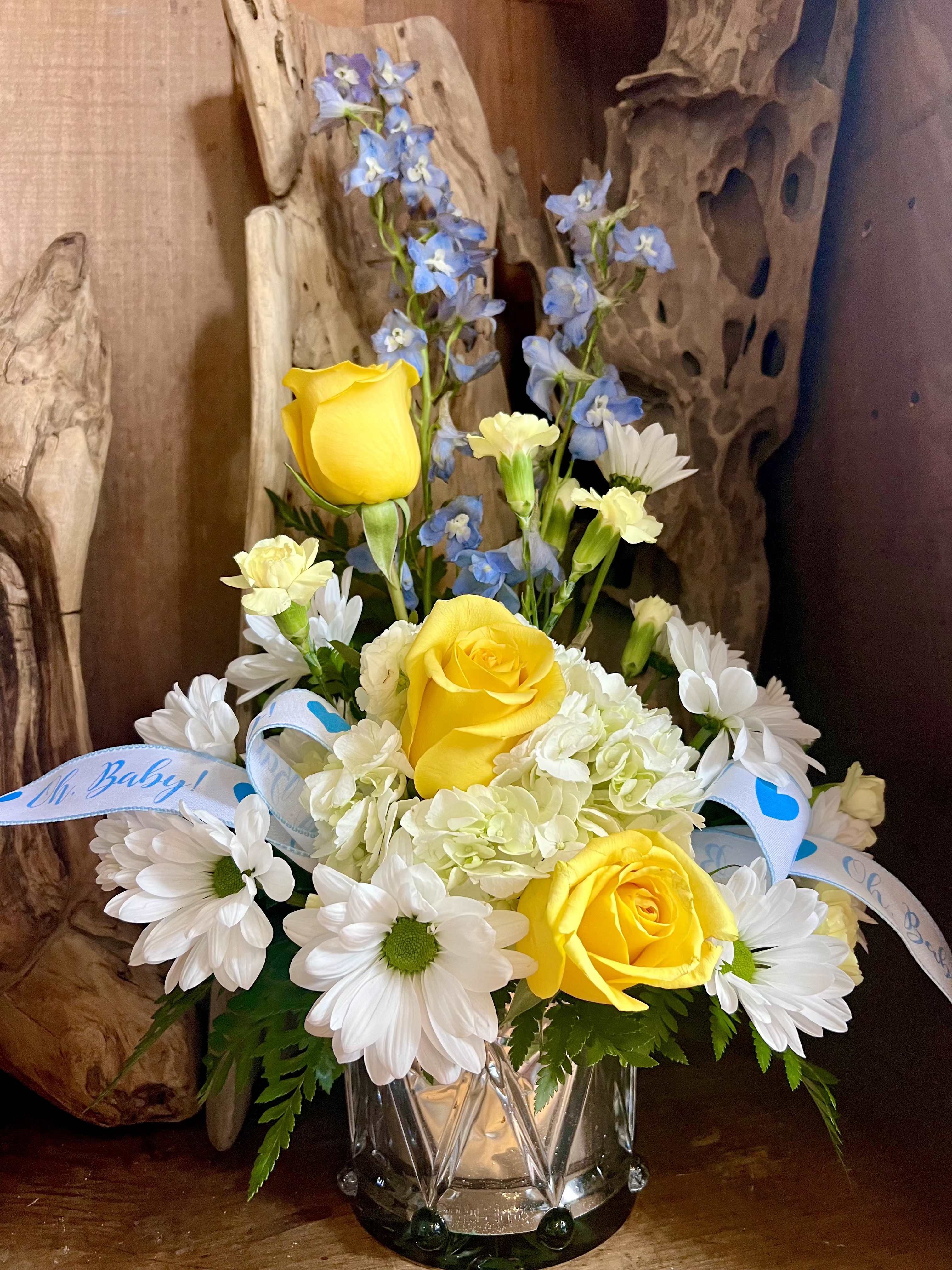 The Little Drummer Boy - Sunny yellow roses meet tall blue delphinium in a joyful, welcoming arrangement perfect for celebrating a new baby, a birthday, or sending a bright "just because" surprise. The Little Drummer Boy flower arrangement places the occasion-forward bouquet where it belongs - the recipient will feel genuinely celebrated by the cheerful palette and thoughtful nursery-ready accents.  Clear faceted glass holds a full, upright composition of yellow roses, white daisies, pale green hydrangea, blue delphinium spikes, and soft carnations. The delphinium rises above the rounder blooms to give height and movement, while the daisies and carnations add friendly texture. A light blue decorative ribbon with baby-themed text ties the design together and reinforces the gentle, welcoming mood.  Ideal for placing on a nursery dresser, kitchen table, or office desk, this arrangement reads as both celebratory and comfortingly familiar - the kind of gift that makes someone feel truly seen. Arrives in a glass vase ready to display; refresh water every two to three days and trim stems as needed to keep blooms looking their best.