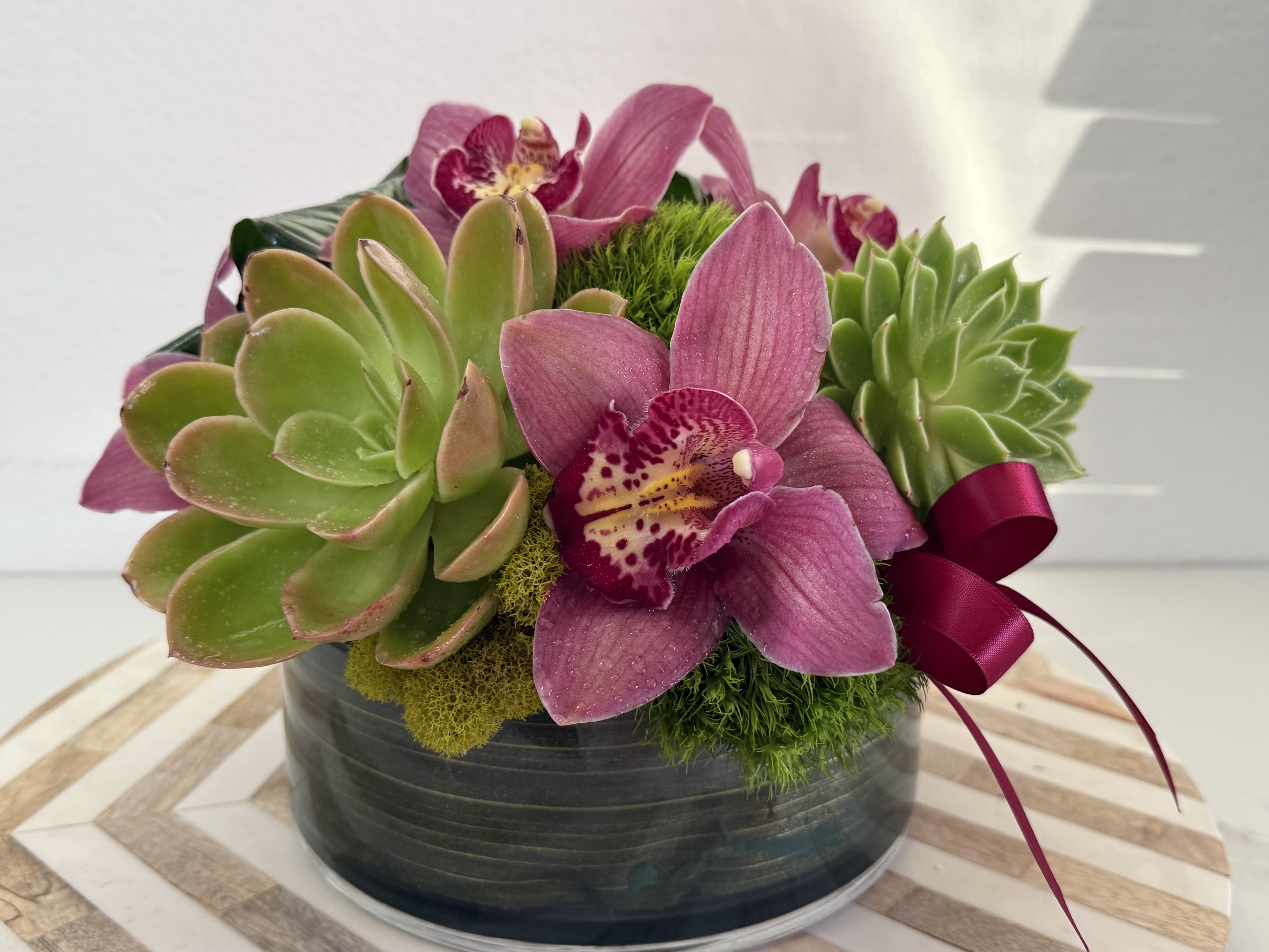 Pink orchids and green succulents arranged in a round black container with a ribbon